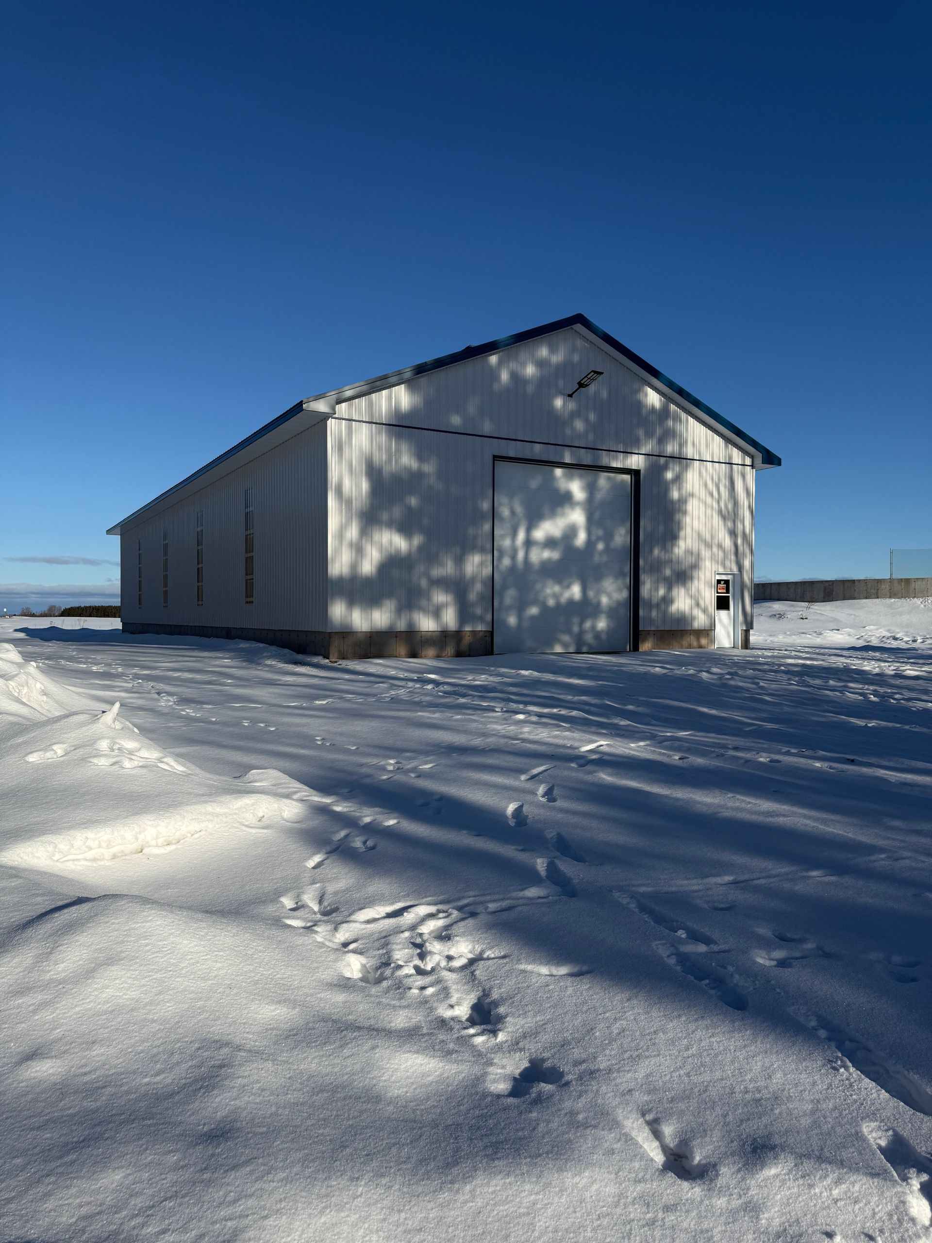 Une grange blanche se dresse au milieu d'un champ enneigé, sous un ciel d'un bleu limpide, les ombres des arbres se projetant sur le bâtiment.
