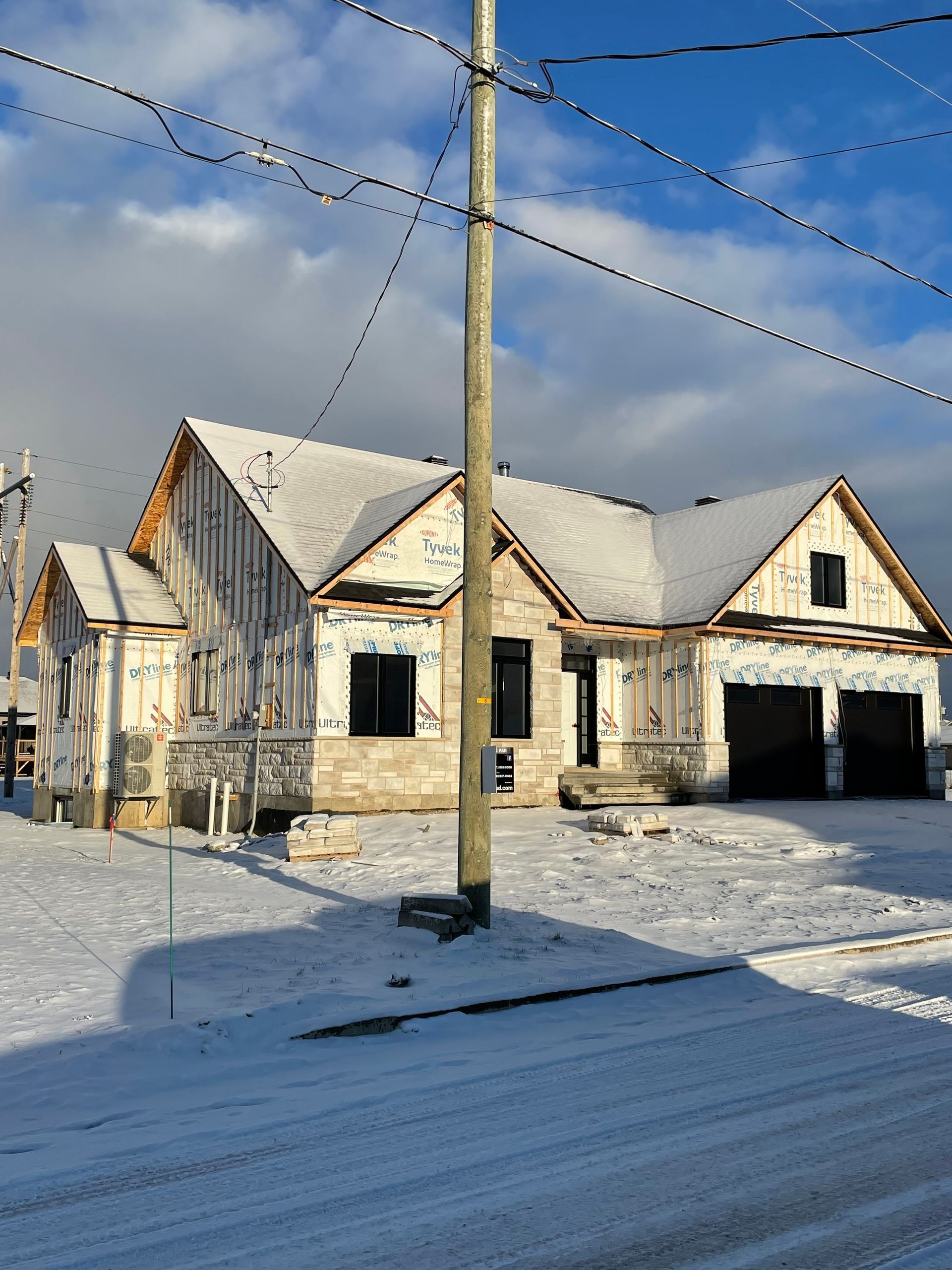 Une maison de plain-pied en construction, avec un bardage blanc et des portes de garage noires, entourée de neige sous un ciel bleu.