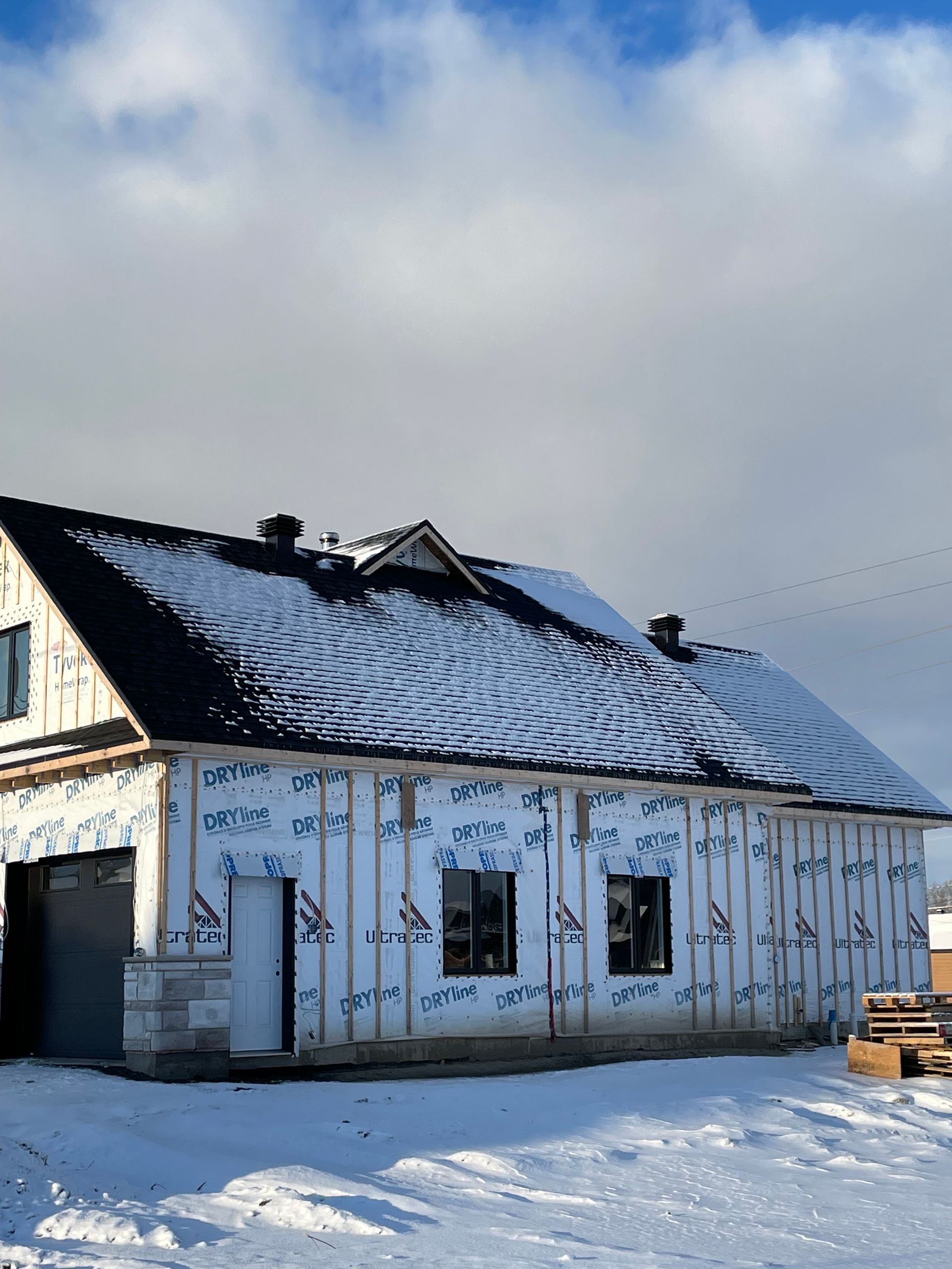 Une maison neuve en construction, avec un toit enneigé, des fenêtres sombres et une façade blanche se détachant sur un ciel bleu.