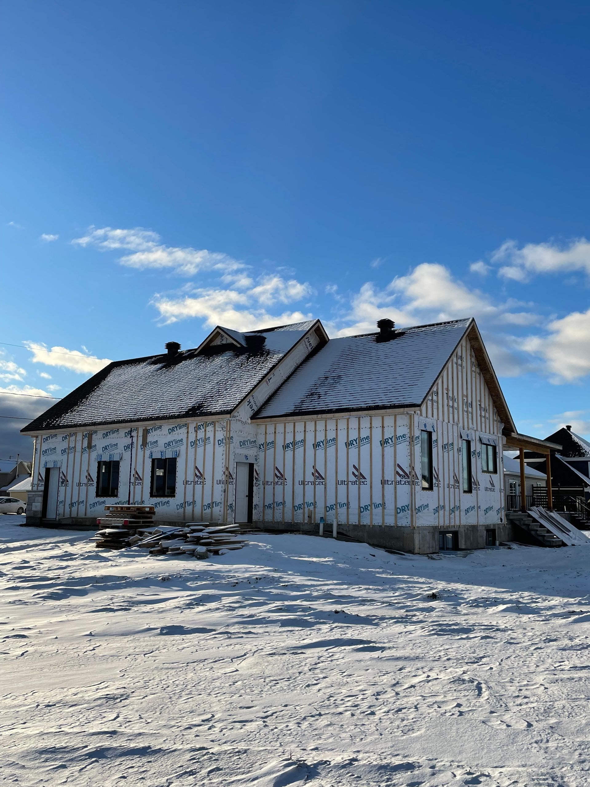Une maison en construction, recouverte d'une bâche de protection blanche, se dresse dans un champ enneigé sous un ciel bleu azur.