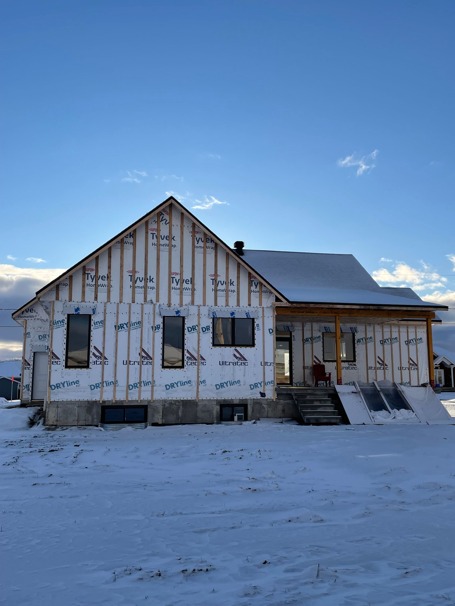 Une maison en construction, avec un revêtement extérieur blanc, une charpente en bois et un sol enneigé sous un ciel bleu.