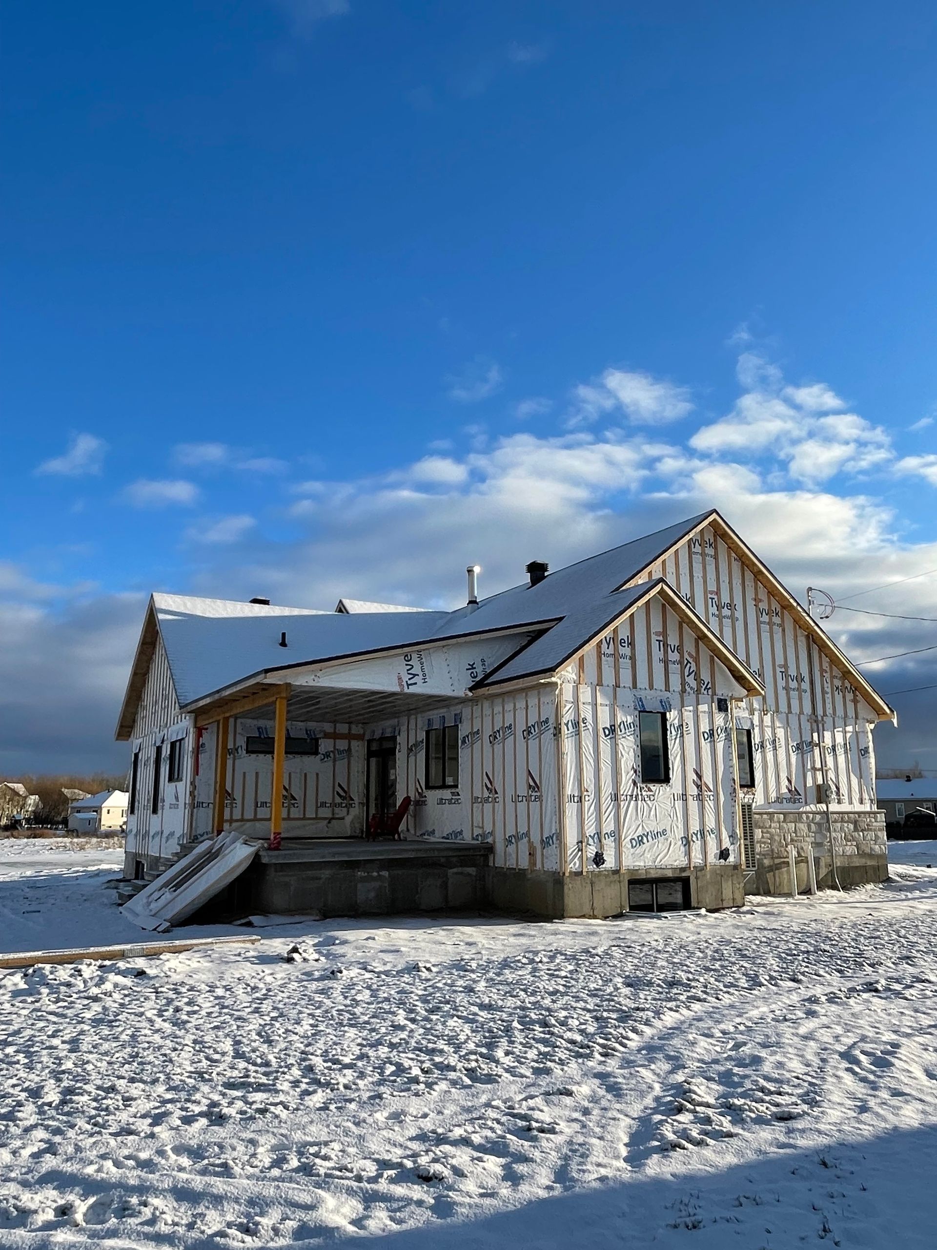 Une maison en construction, recouverte de bardage blanc, se dresse au milieu d'un champ enneigé sous un ciel d'un bleu éclatant.