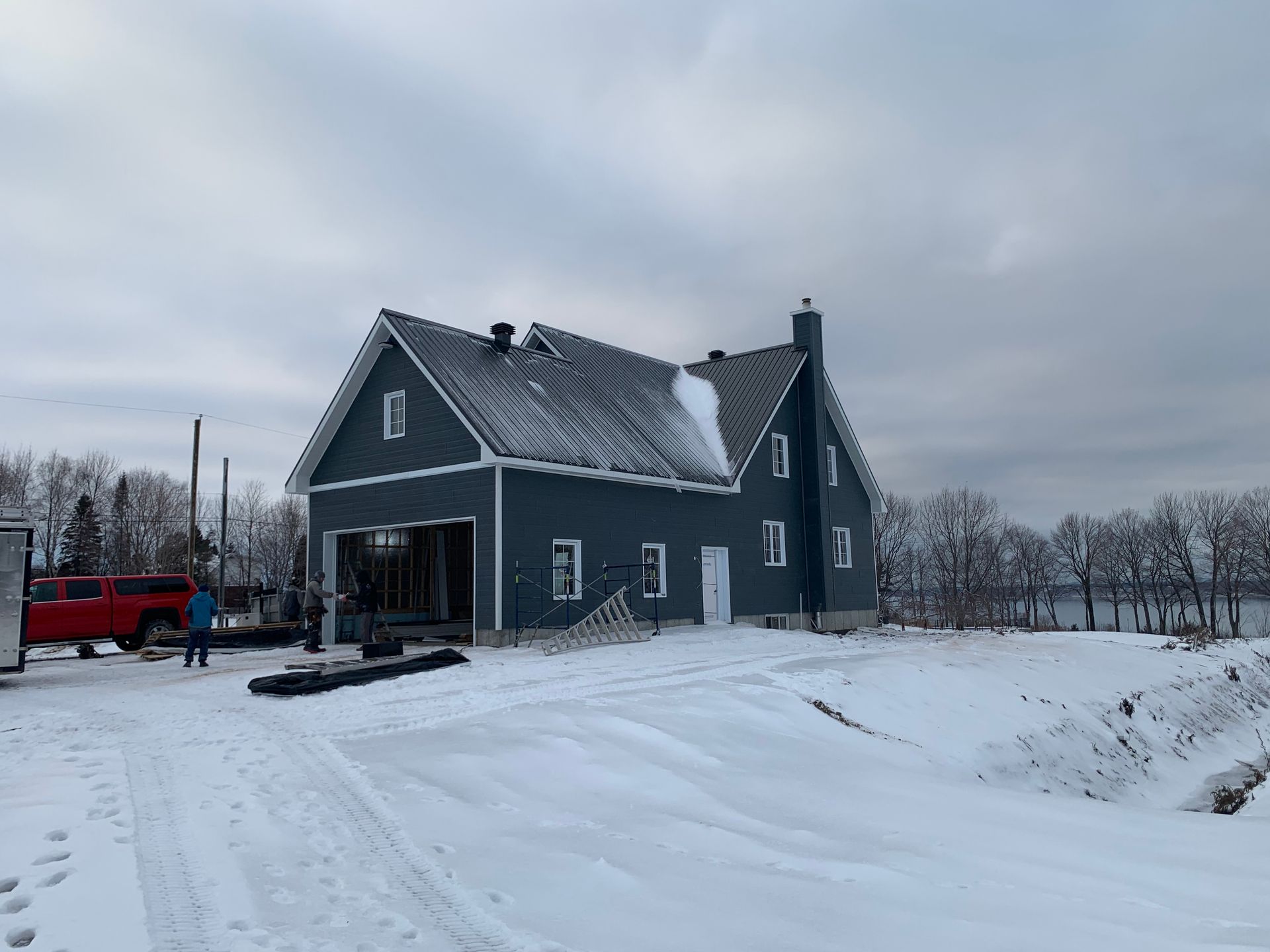 Une maison gris foncé à deux étages, avec un toit en métal, en construction, entourée de neige, avec un camion rouge garé à proximité.