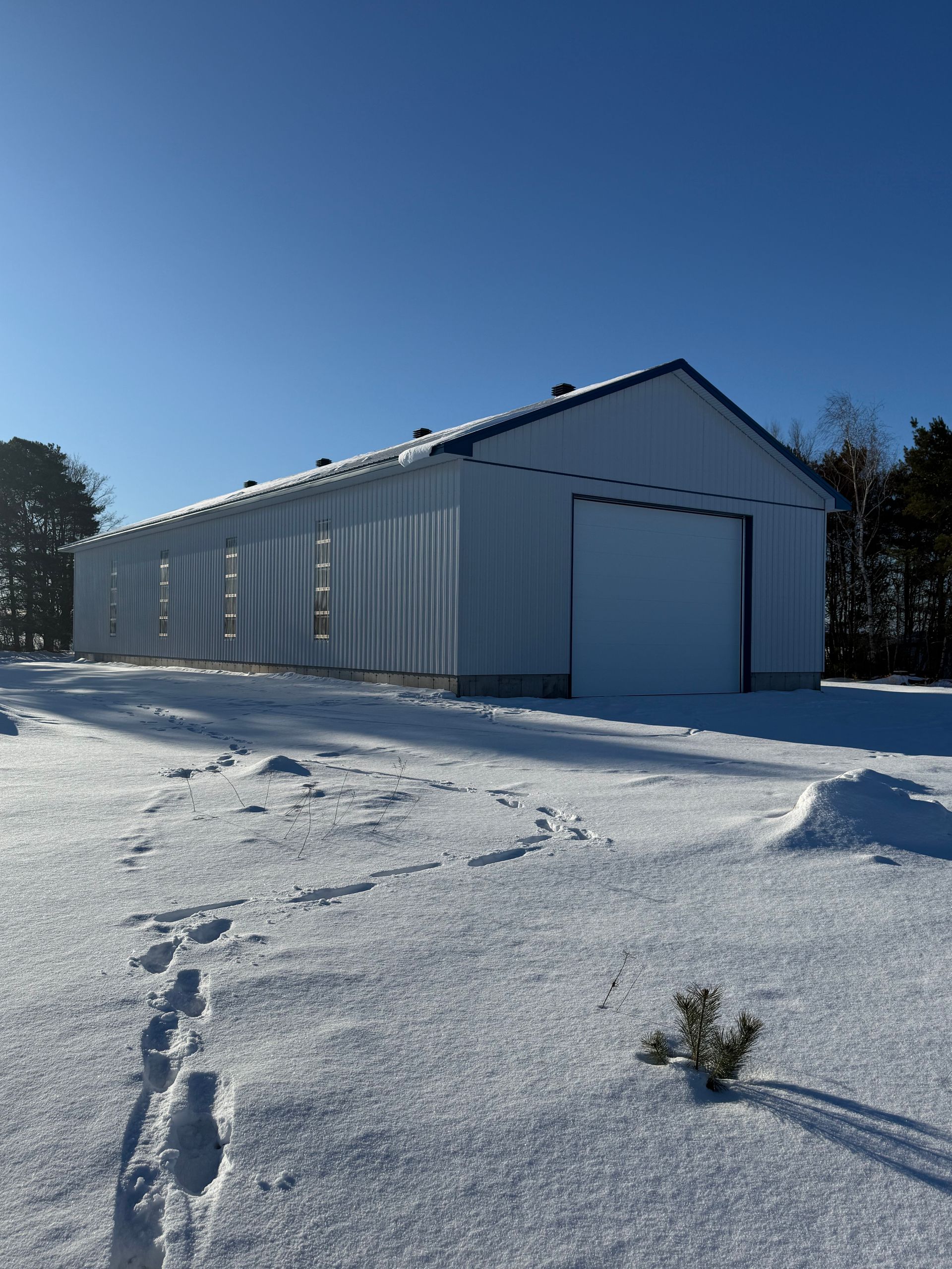 Un bâtiment en métal blanc se dresse au milieu d'un champ enneigé, sous un ciel d'un bleu limpide, un chemin d'empreintes de pas y menant.