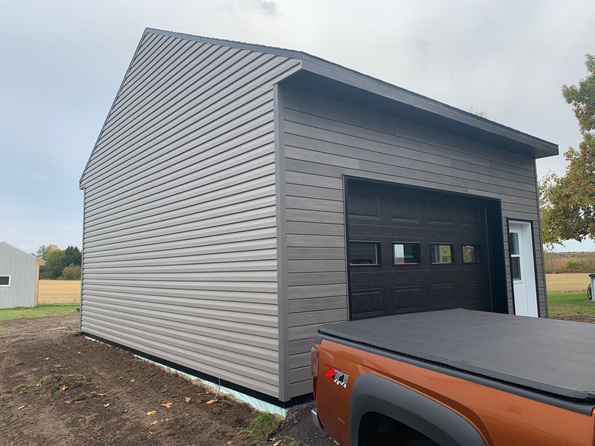 Un garage gris nouvellement construit, avec une porte sombre, se dresse sur un terrain en terre battue, partiellement masqué par la benne d'un camion orange.