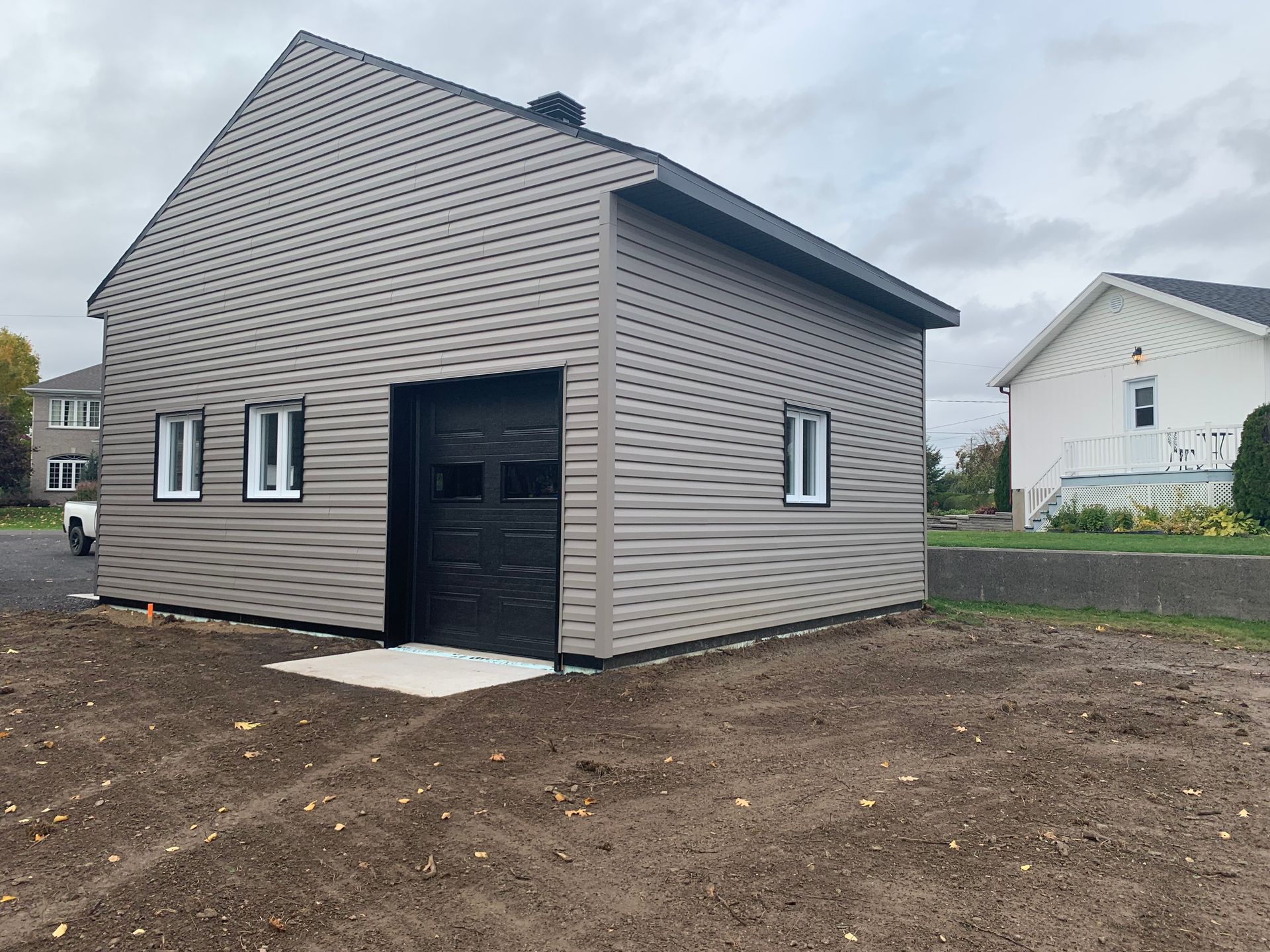 Un garage indépendant nouvellement construit, avec un bardage horizontal brun clair, une porte de garage noire et des fenêtres à encadrement blanc.