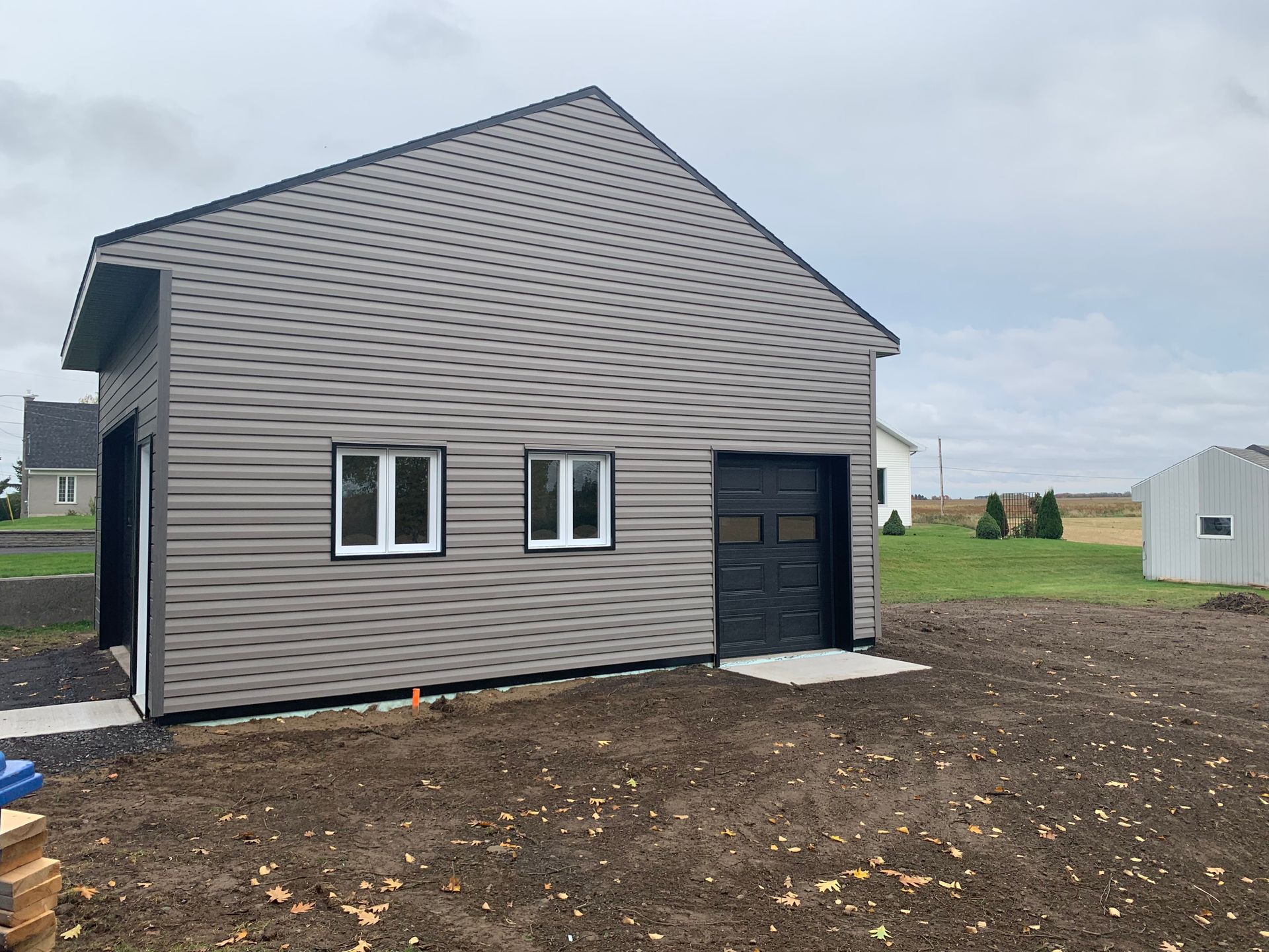Un garage gris, nouvellement construit, avec des parois métalliques, des portes noires et deux fenêtres, situé sur un terrain en terre battue non aménagé.
