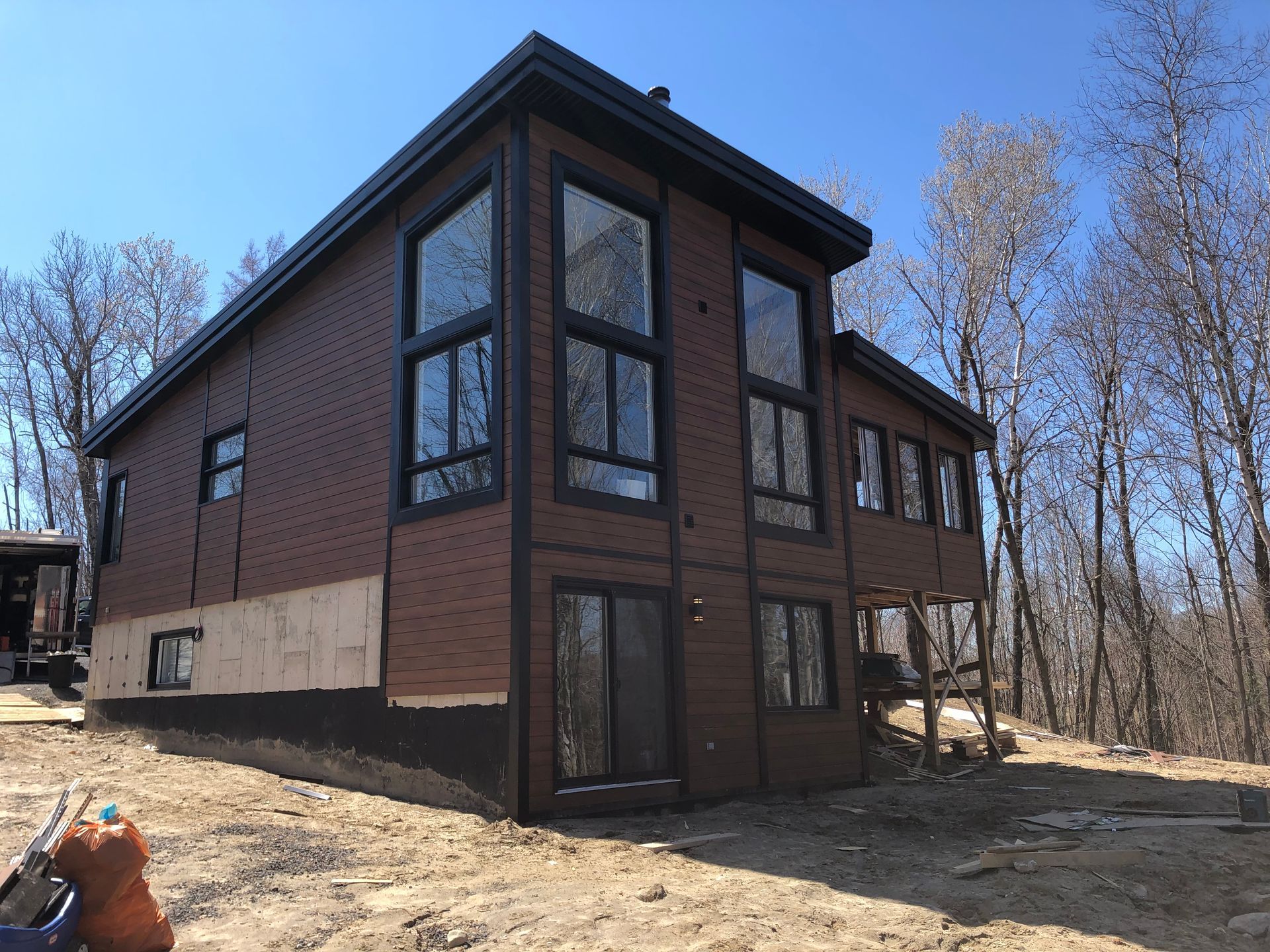 Une maison de deux étages, de couleur brun foncé et dotée de grandes fenêtres, se dresse sur un terrain boisé en construction, sous un ciel d'un bleu limpide.