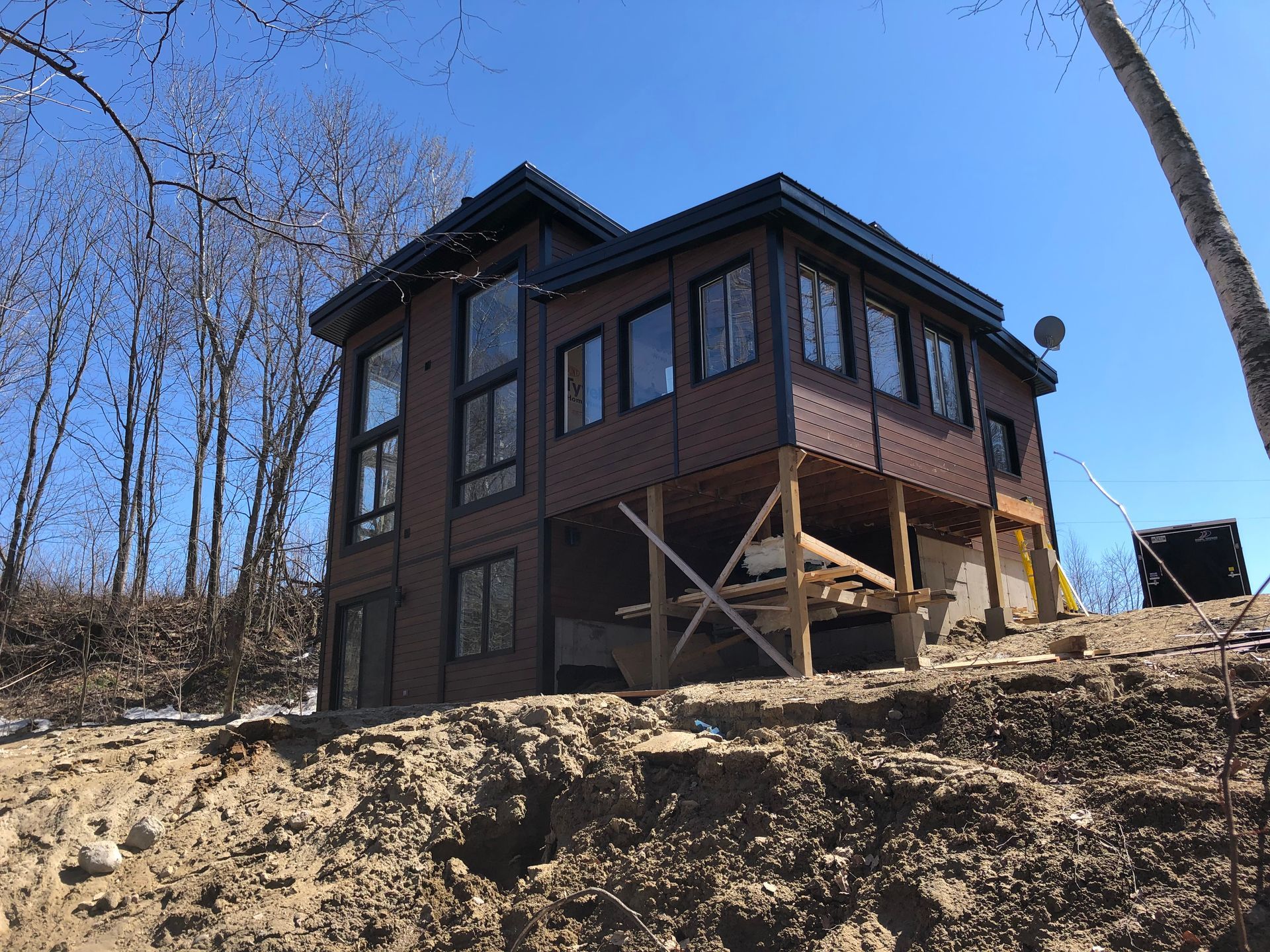 Une maison moderne brune aux grandes fenêtres se dresse sur un flanc de colline en pente, partiellement déboisé, sous un ciel d'un bleu éclatant.