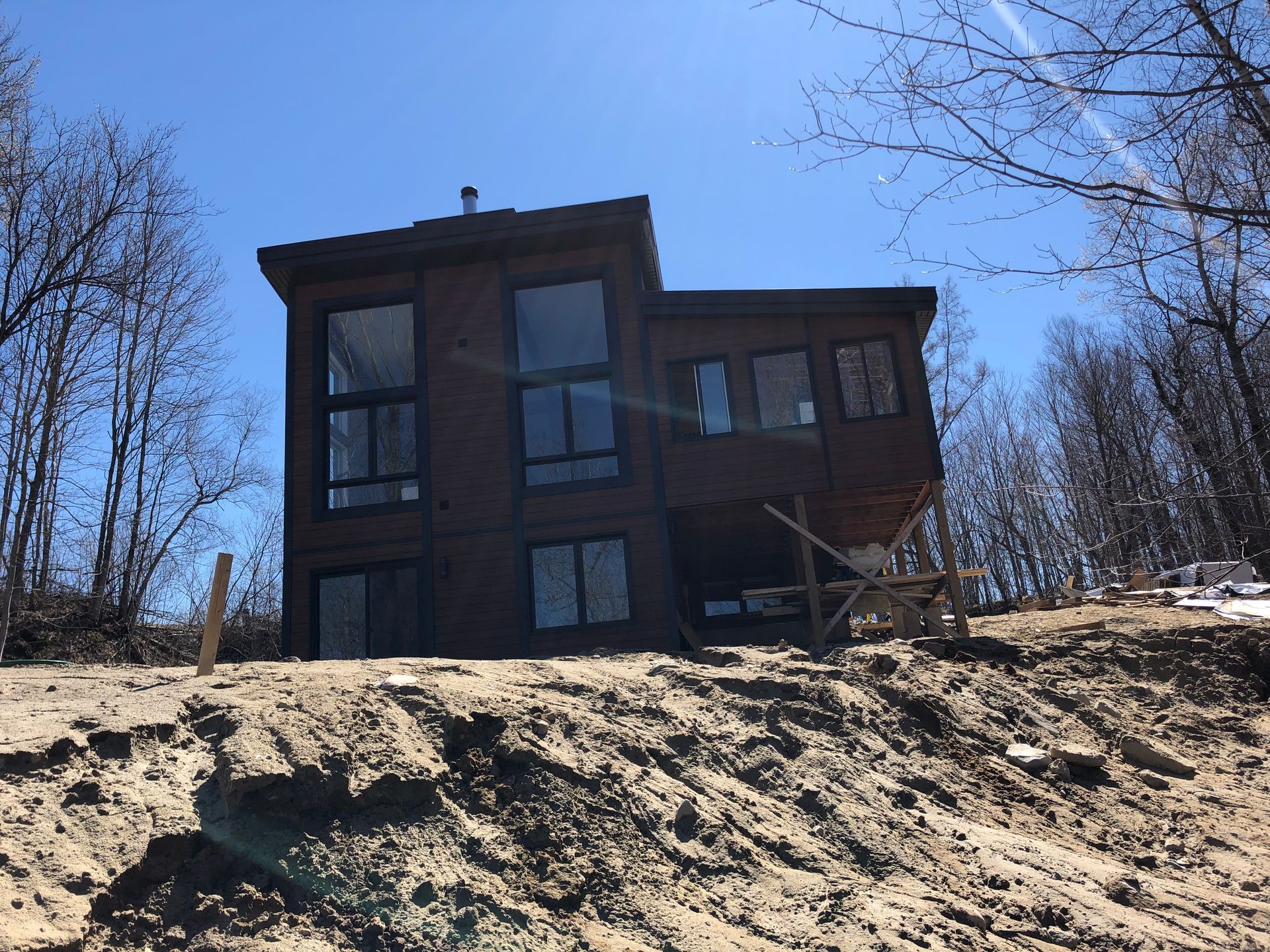 Une maison moderne aux tons sombres et aux grandes fenêtres se dresse au sommet d'une colline sablonneuse en pente, sous un ciel d'un bleu clair et lumineux.