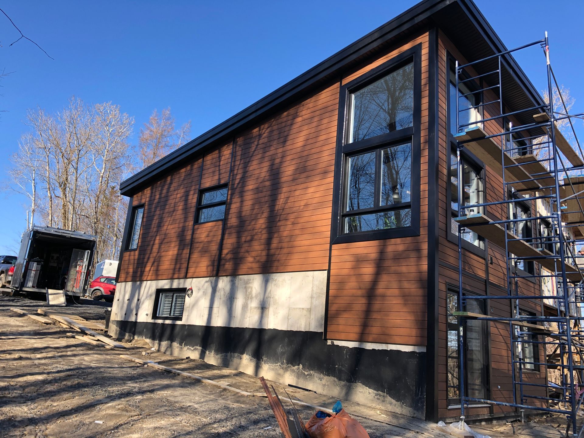 Une maison en construction, avec un bardage marron, de grandes fenêtres et des échafaudages sur le côté, située sur un terrain en pente.