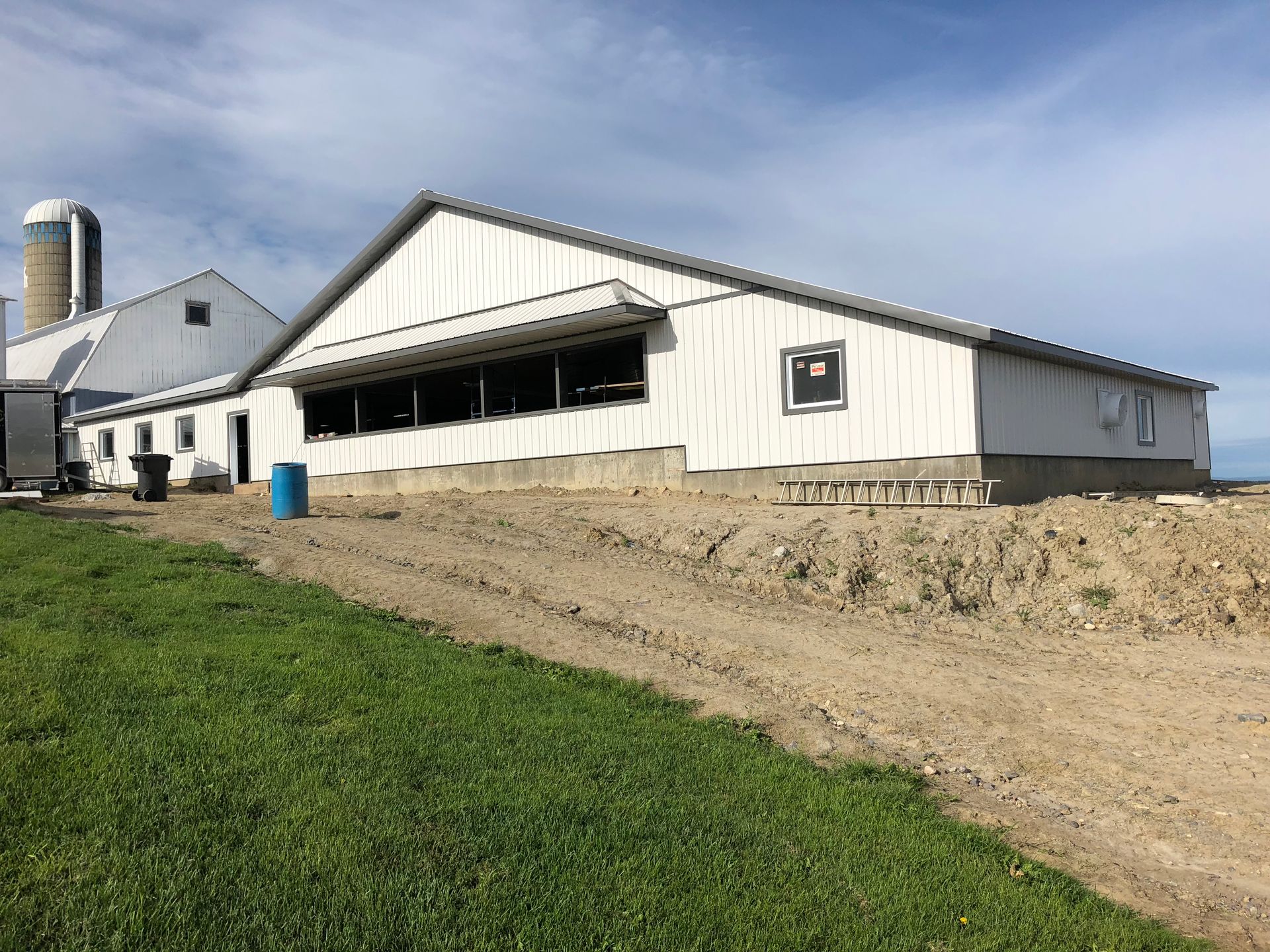 Une longue grange agricole blanche sous un ciel bleu, avec un chemin de terre au premier plan et un silo à l'arrière-plan.