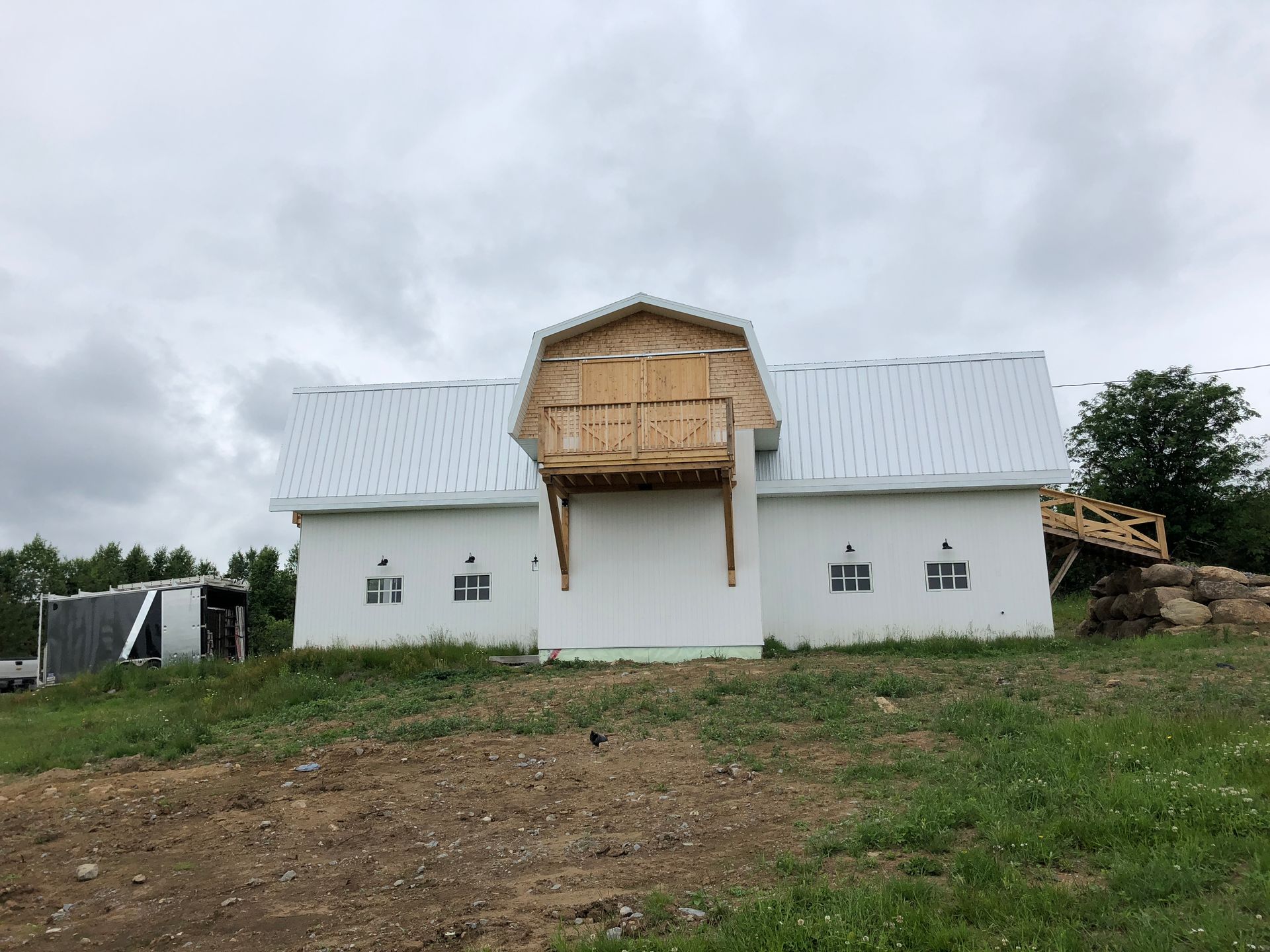 Une grange blanche au toit métallique possède une porte-fenêtre centrale en bois surélevée donnant sur un grenier et un balcon, le tout dans un champ herbeux.