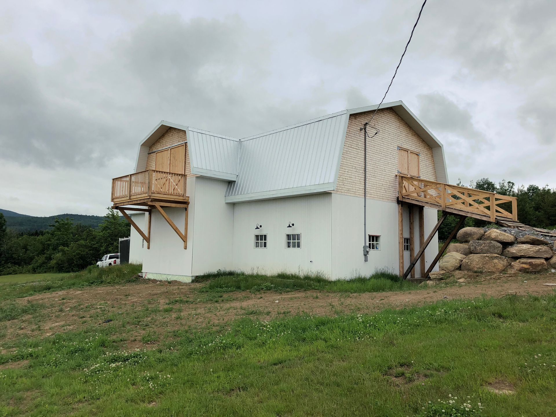 Une grange blanche au toit à deux pans brisés, deux balcons en bois et une rampe en terre menant à une porte à l'étage, donnant sur un champ herbeux.