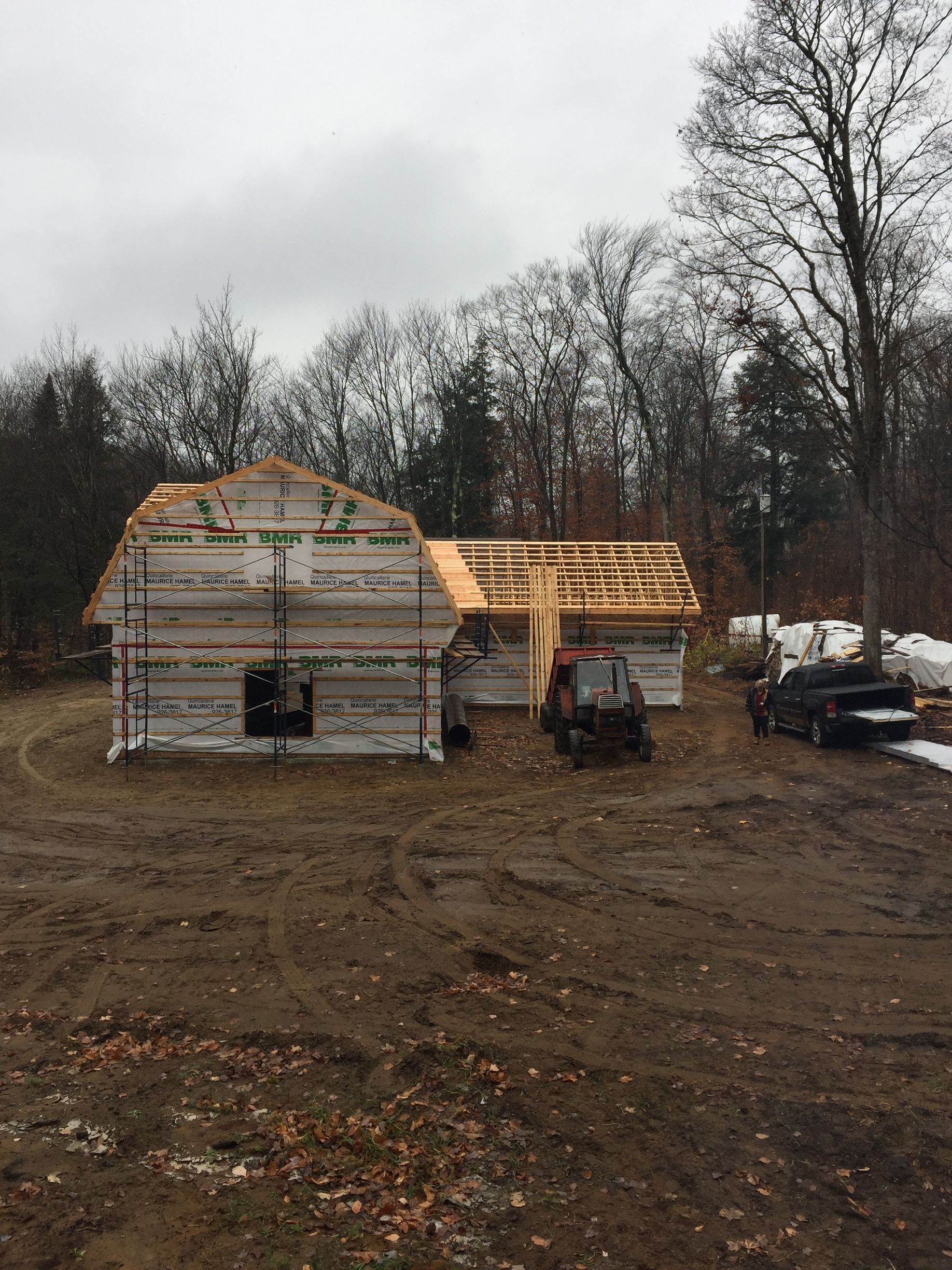 Une maison à moitié construite, avec des panneaux isolants blancs, sous un ciel nuageux, sur un terrain boisé et boueux, avec un camion à proximité.