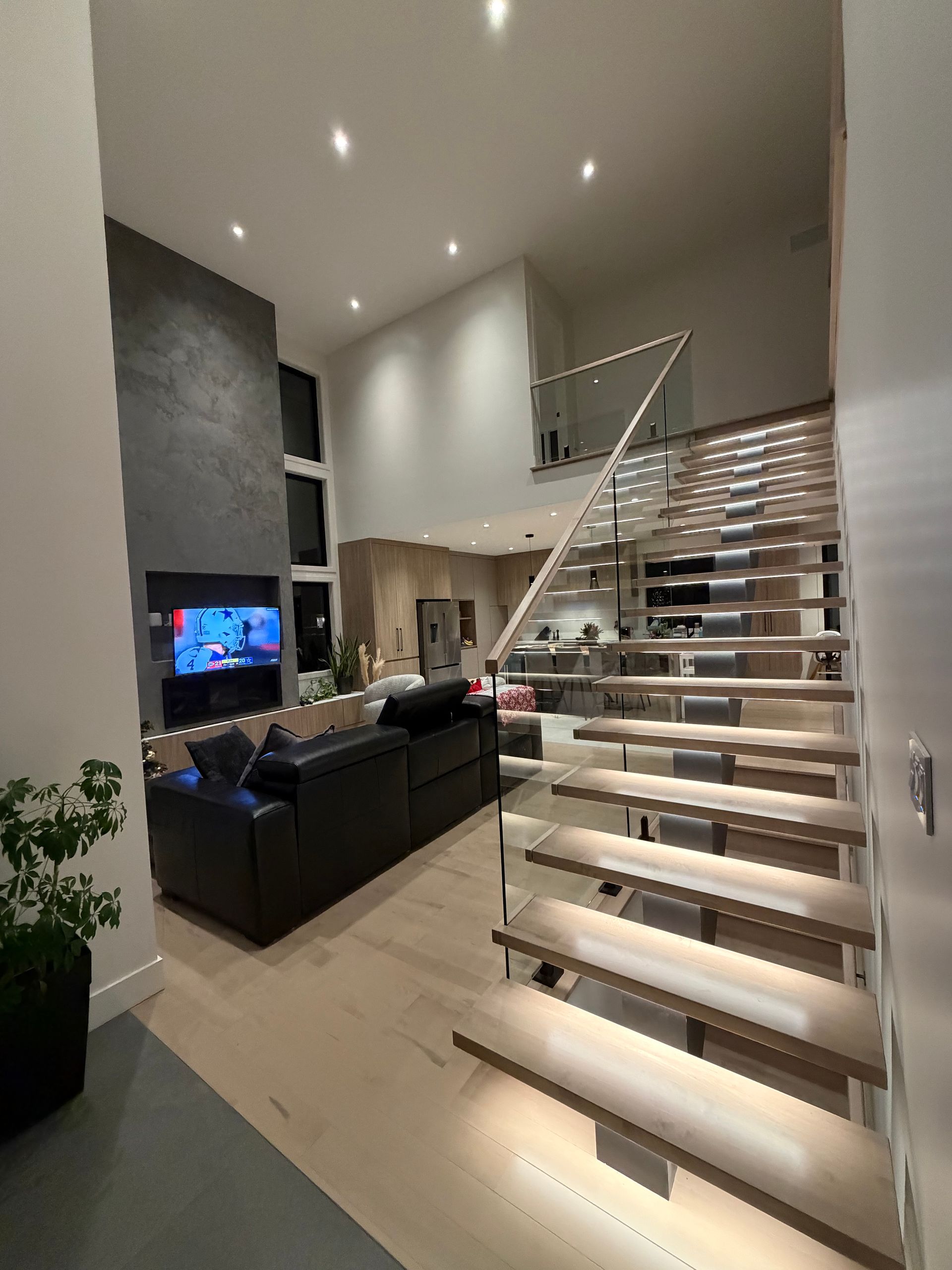 Un salon moderne et ouvert avec une cheminée sombre, un canapé noir et un escalier éclairé avec une rampe en verre.