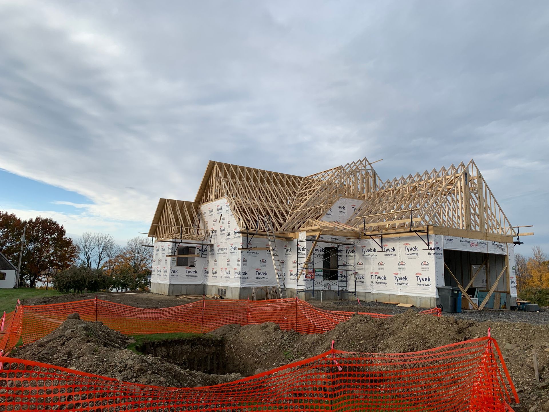 Une maison en construction avec une charpente apparente en bois et un revêtement extérieur blanc, entourée d'un grillage de sécurité orange.