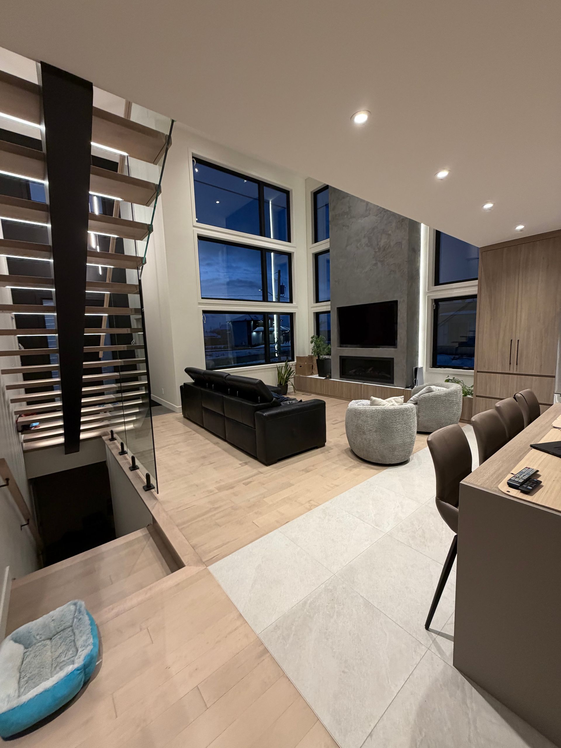 Un salon moderne à aire ouverte avec parquet clair, un canapé noir, deux fauteuils gris et un escalier à côté de grandes fenêtres.