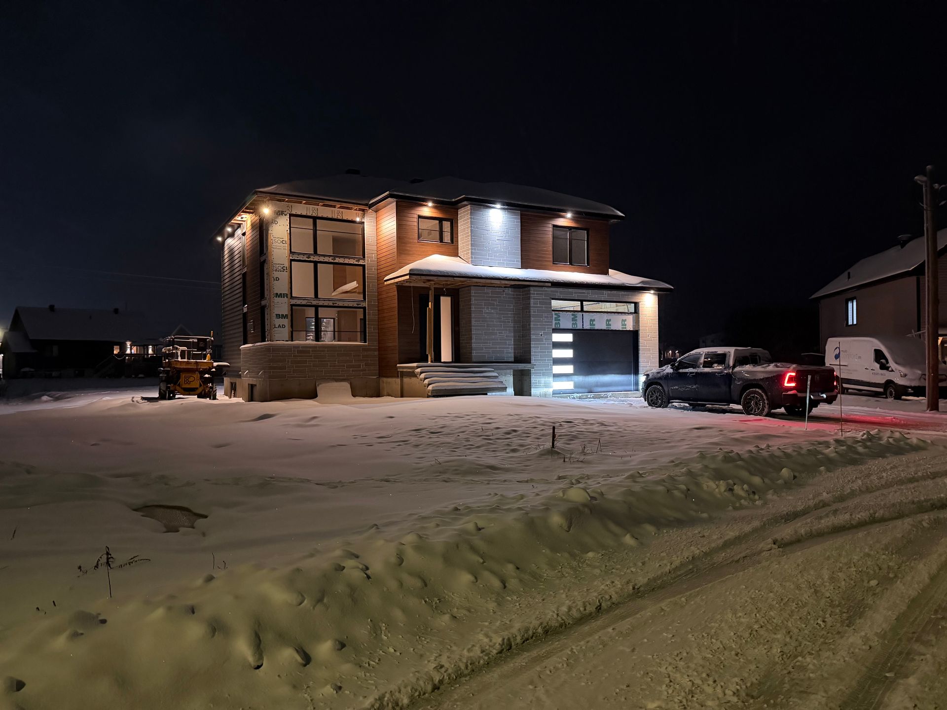 Une maison moderne de deux étages en construction, la nuit, éclairée par des projecteurs extérieurs, sur un terrain enneigé avec un camion.