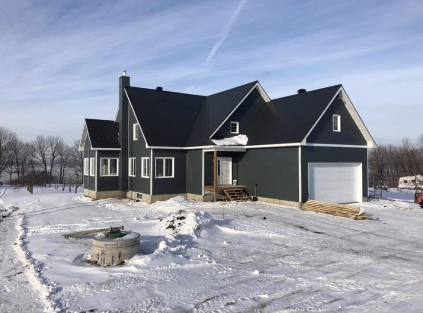 Une maison gris foncé avec un garage et un toit blancs, entourée de neige, sous un ciel bleu clair.