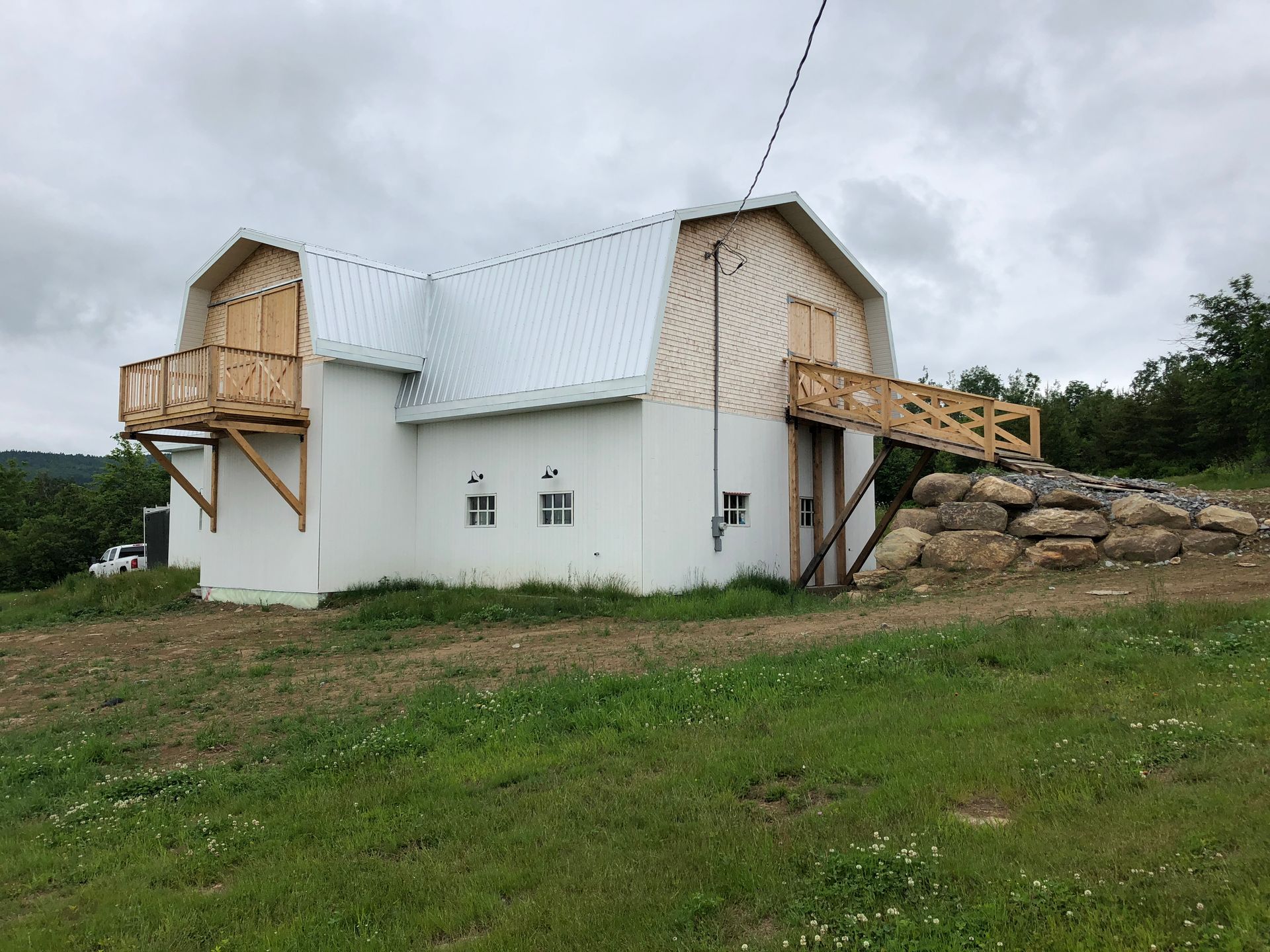 Une grange blanche au toit à deux pans brisés, une terrasse en bois surélevée sur la gauche et un pont en bois menant à un talus rocheux.