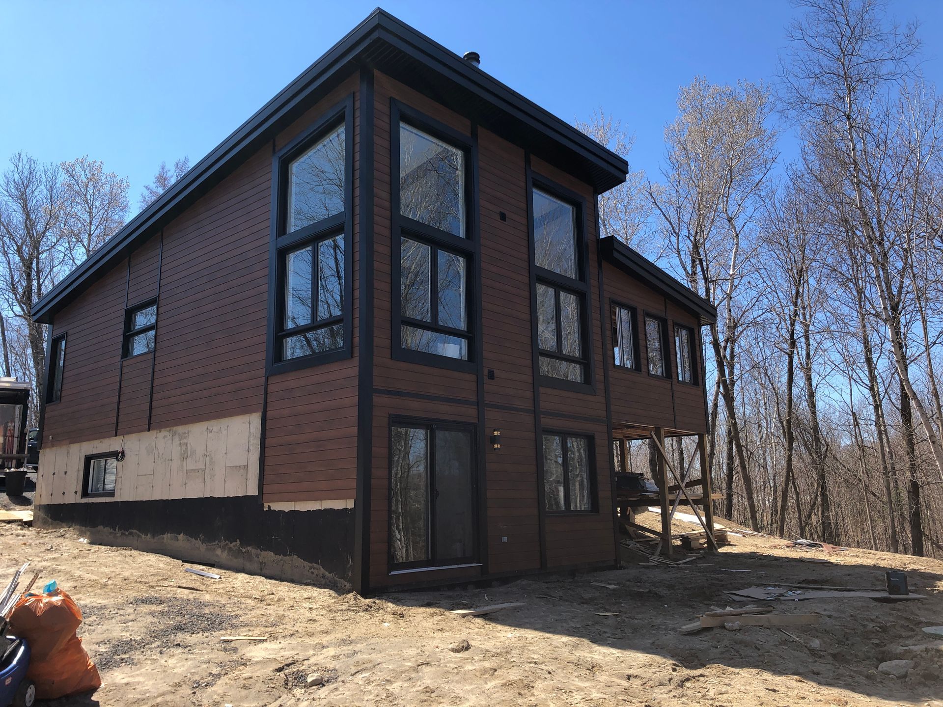 Une maison moderne à deux étages, avec un revêtement en bois brun et de grandes fenêtres, est en construction dans une zone boisée.