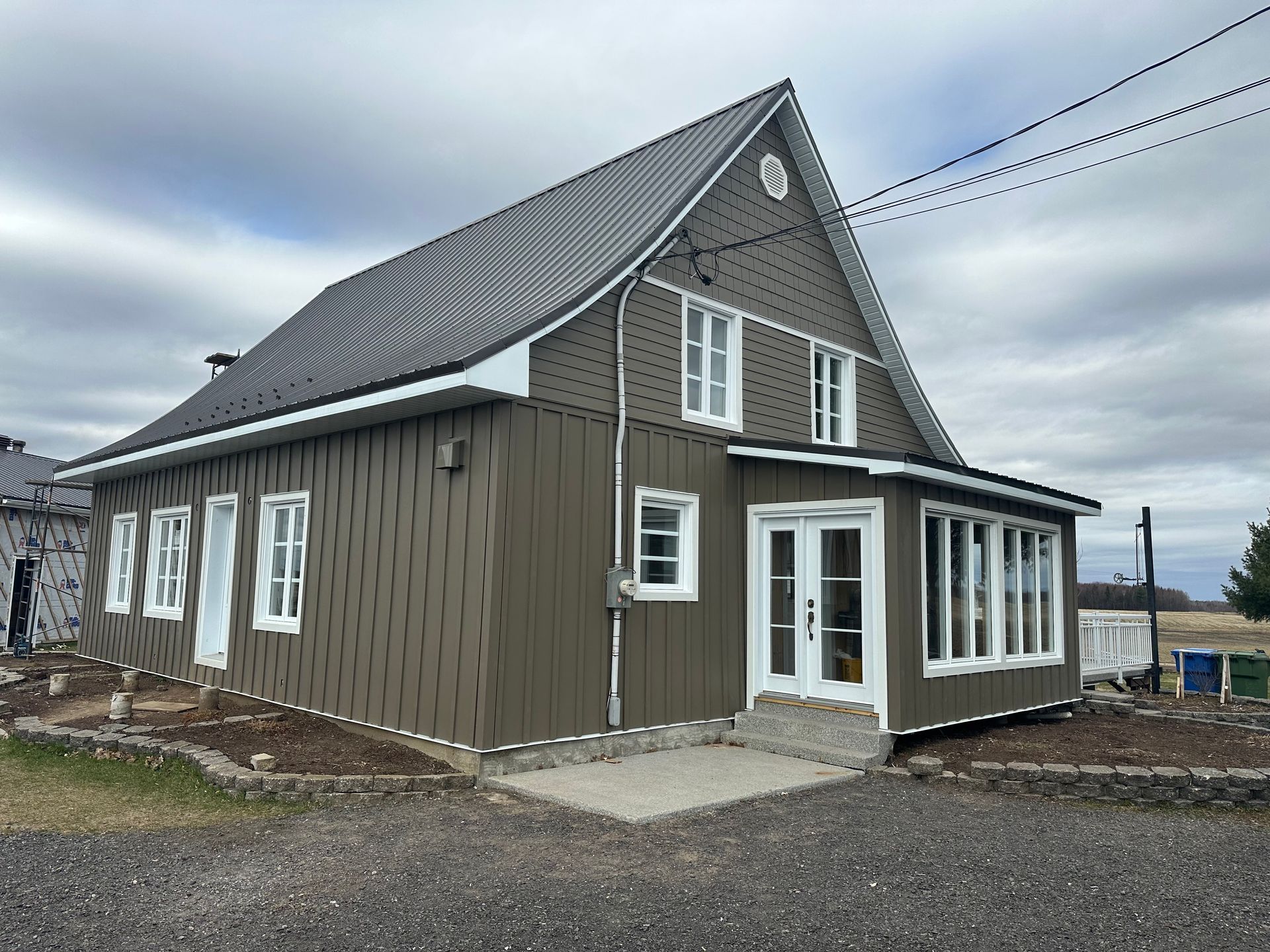 Une maison à deux étages avec un bardage vertical brun foncé, un toit en métal gris et des fenêtres à encadrement blanc, sous un ciel nuageux.