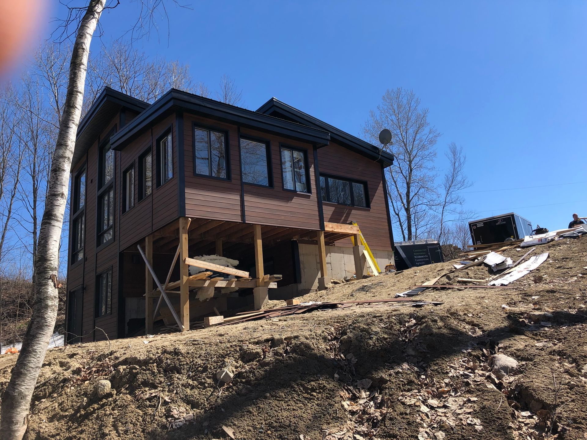 Une maison brune de deux étages en construction, avec une terrasse surélevée, située sur un coteau de terre en pente, sous un ciel bleu azur.