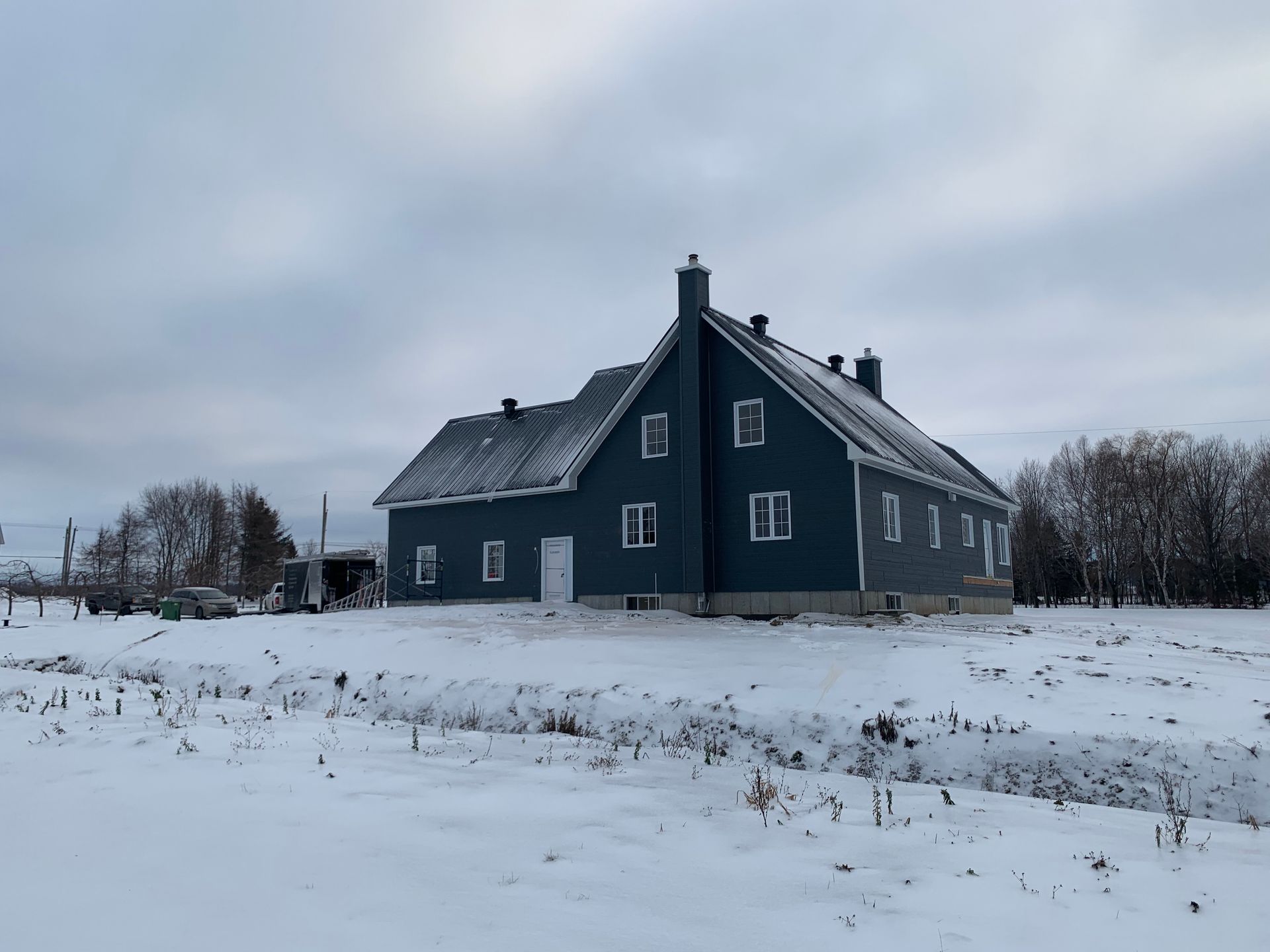 Une maison bleu foncé à deux étages, avec une cheminée proéminente, se dresse dans un paysage plat et enneigé, sous un ciel nuageux.