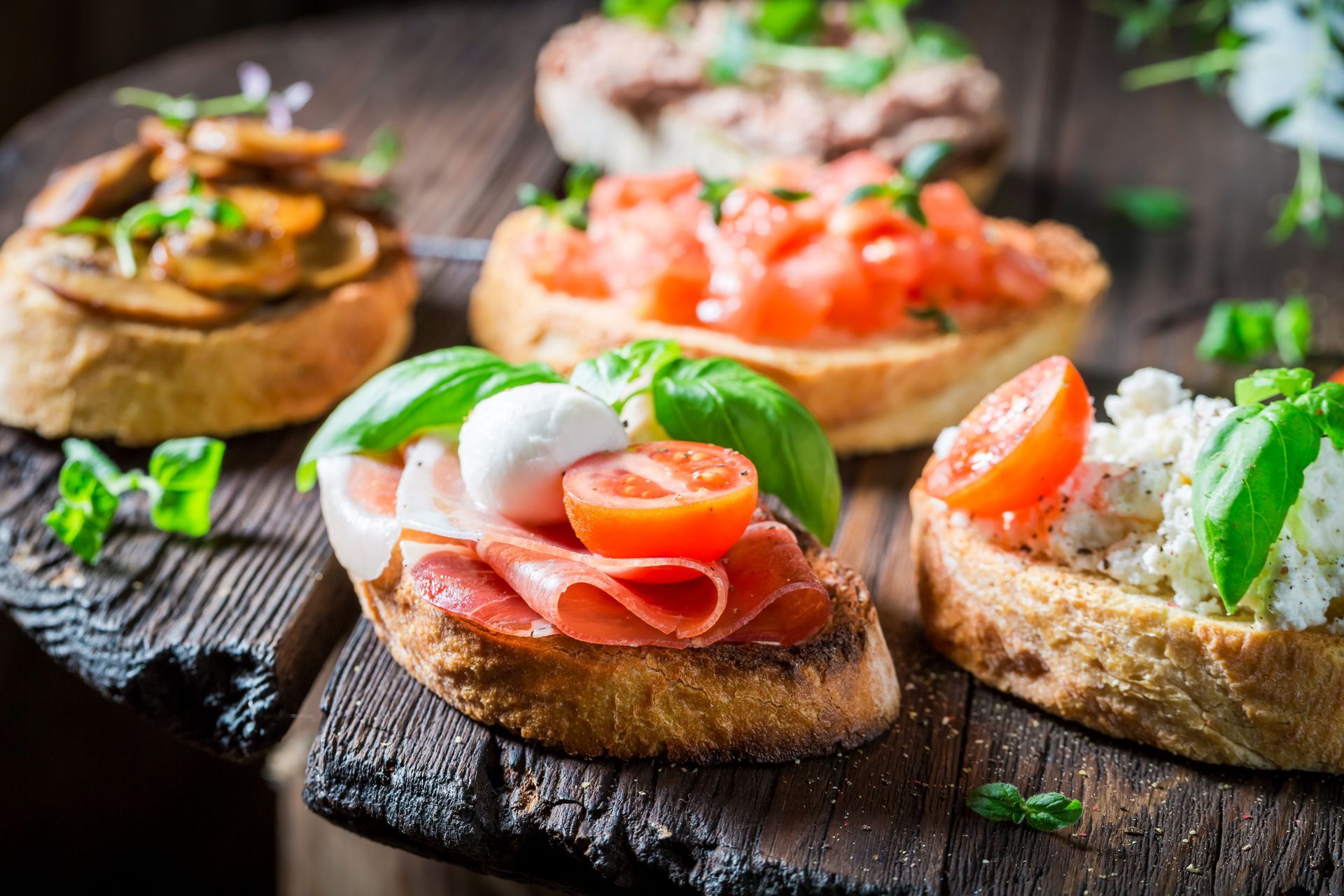 Assorted bruschetta appetizers with tomatoes, basil, and prosciutto on a dark wooden board