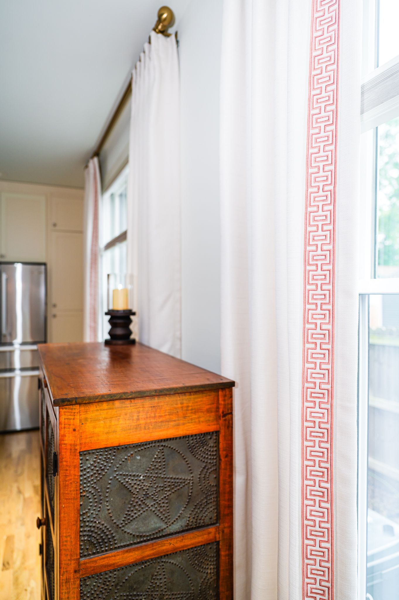 Detail of white drapery with coral Greek-key tape beside a wood pie safe with punched-tin panels near a window.