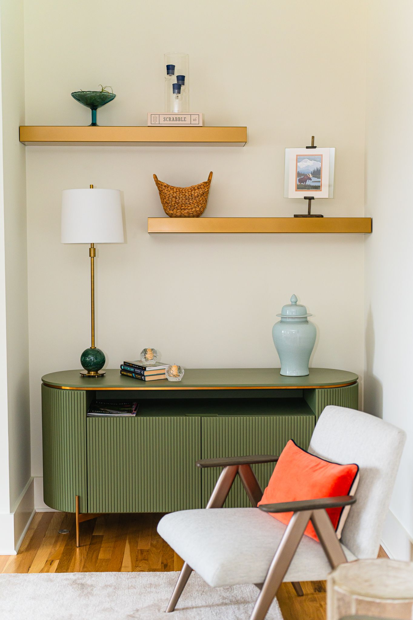 Olive green console with brass shelves and lamp; modern gray chair with coral lumbar pillow.