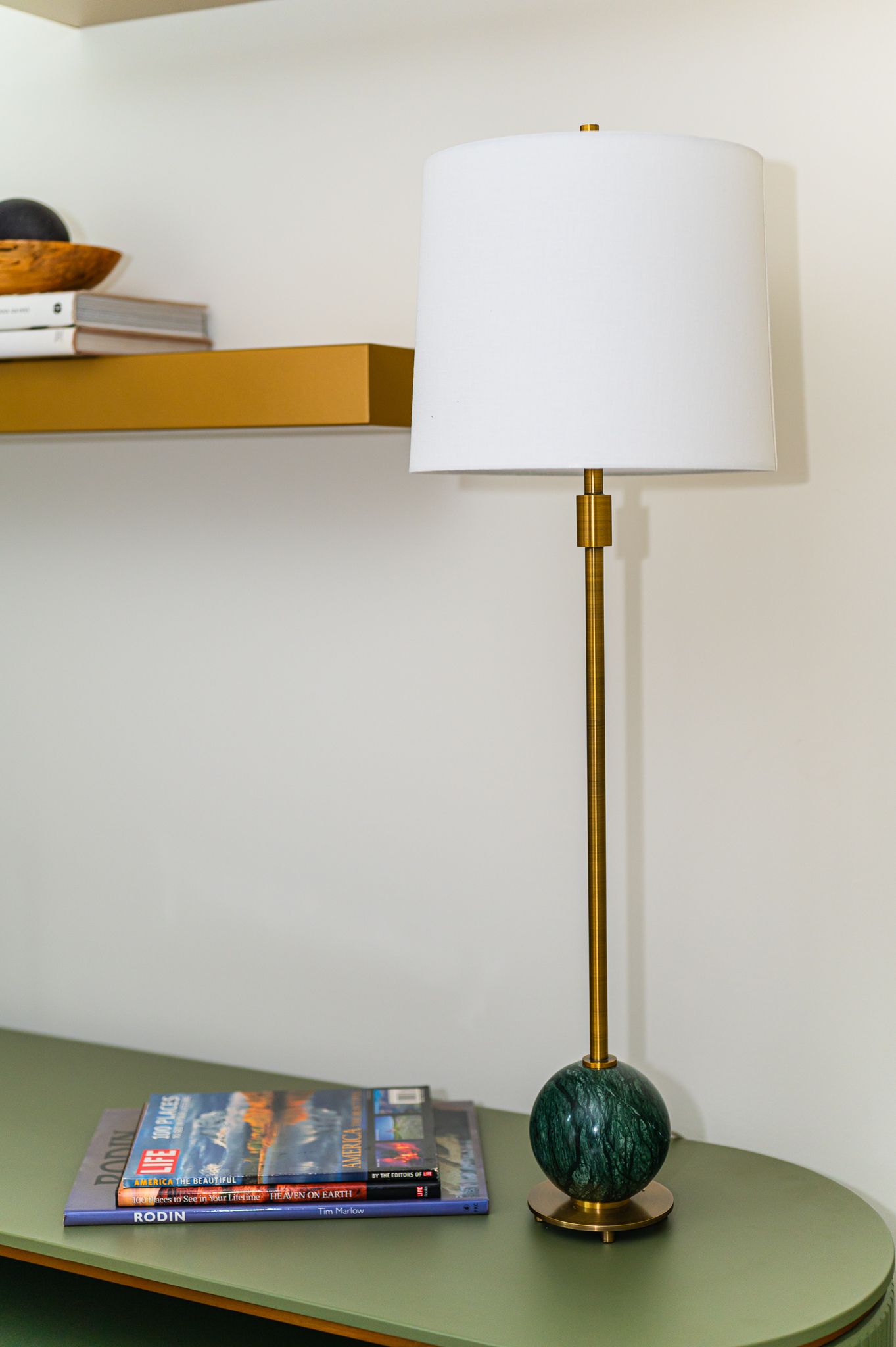 Brass table lamp with white shade and green marble base on olive console under brass floating shelf with books.