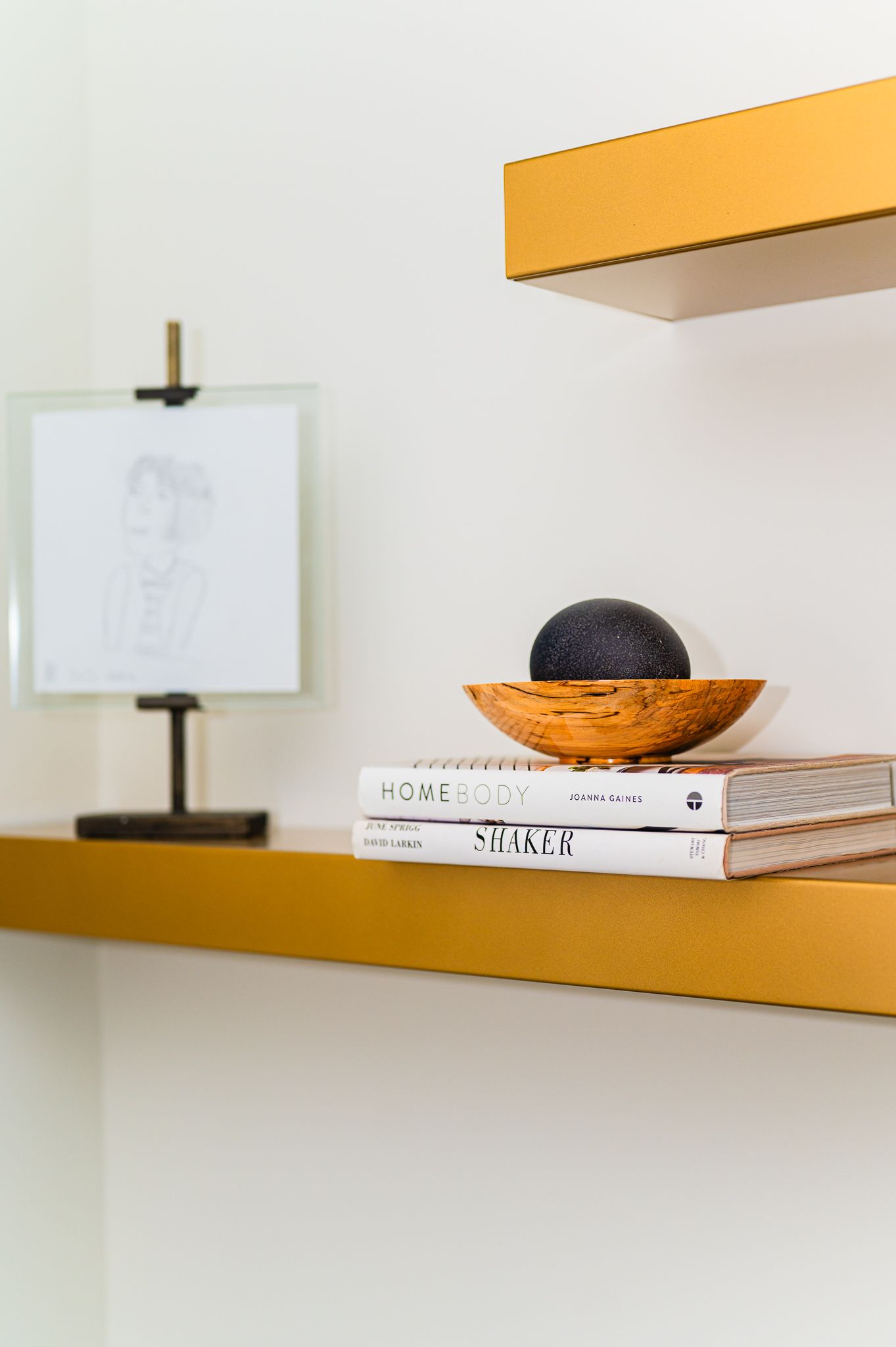 Close-up of brass shelf with stacked books, wood bowl holding a black sphere, and framed sketch in background
