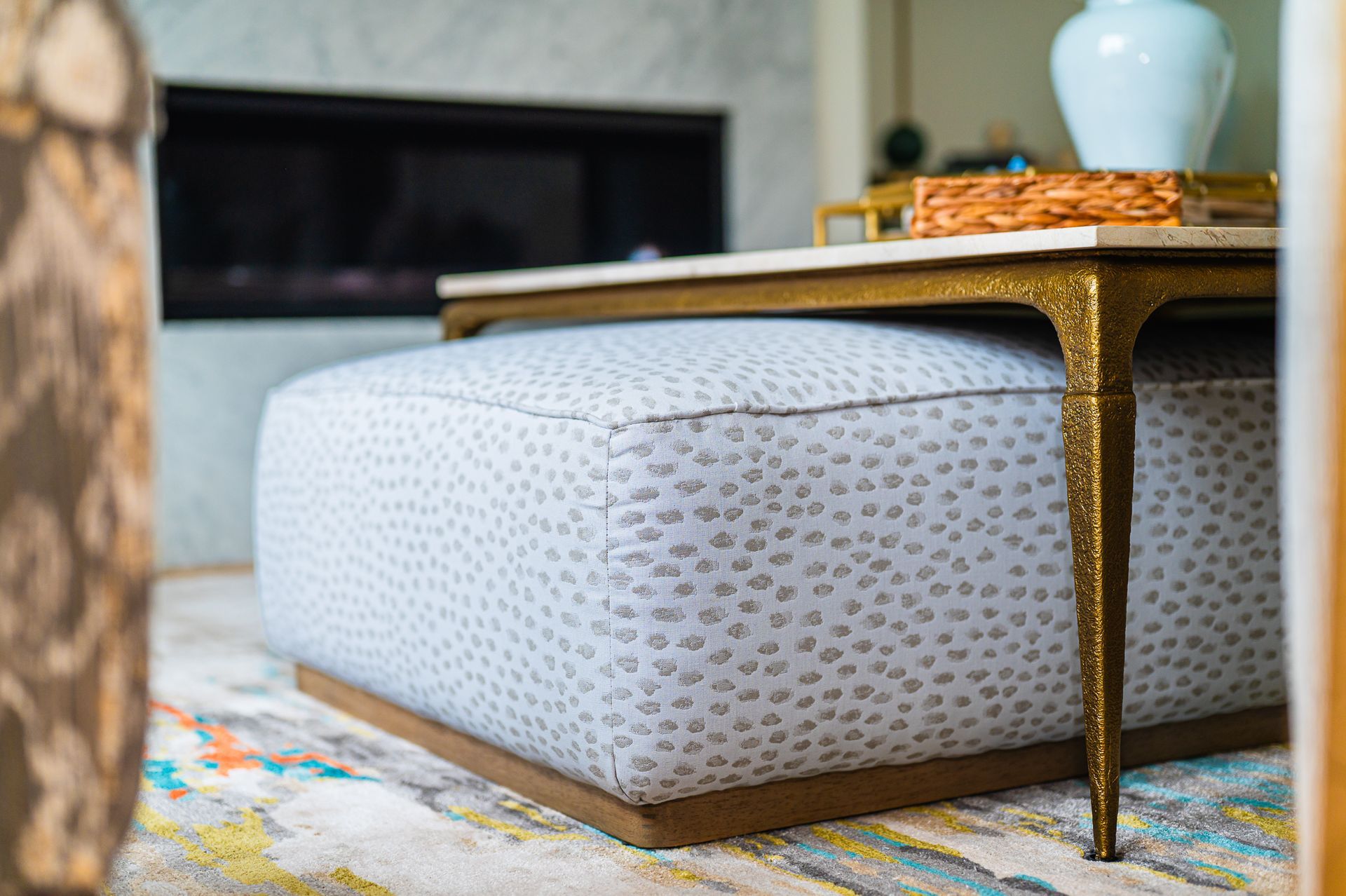 Close-up of dotted light gray ottoman under a brass coffee table leg on a patterned rug.