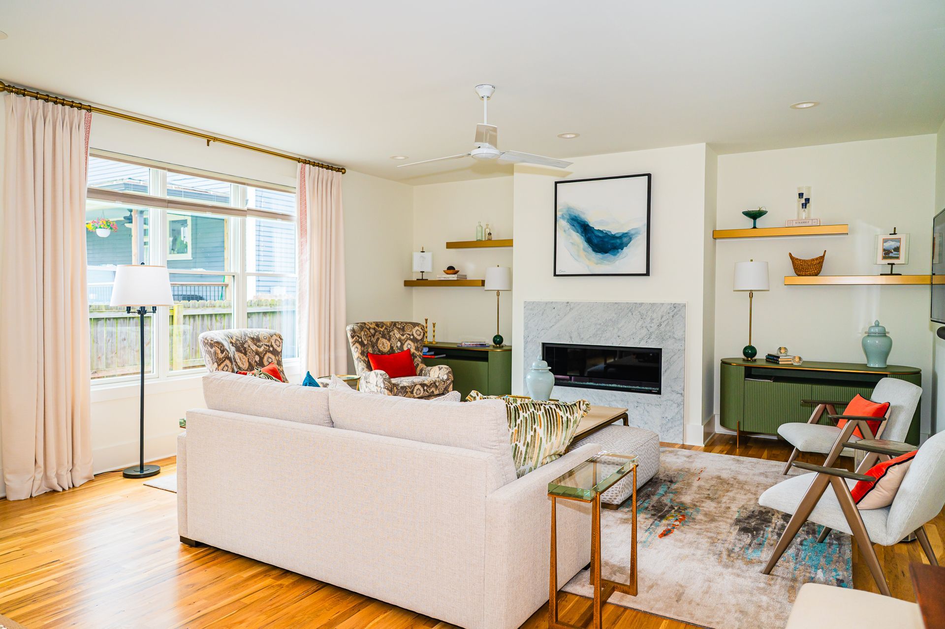 Wide shot of living room with marble fireplace, floating brass shelves, olive consoles, sofa, armchairs, and large window with light drapery.
