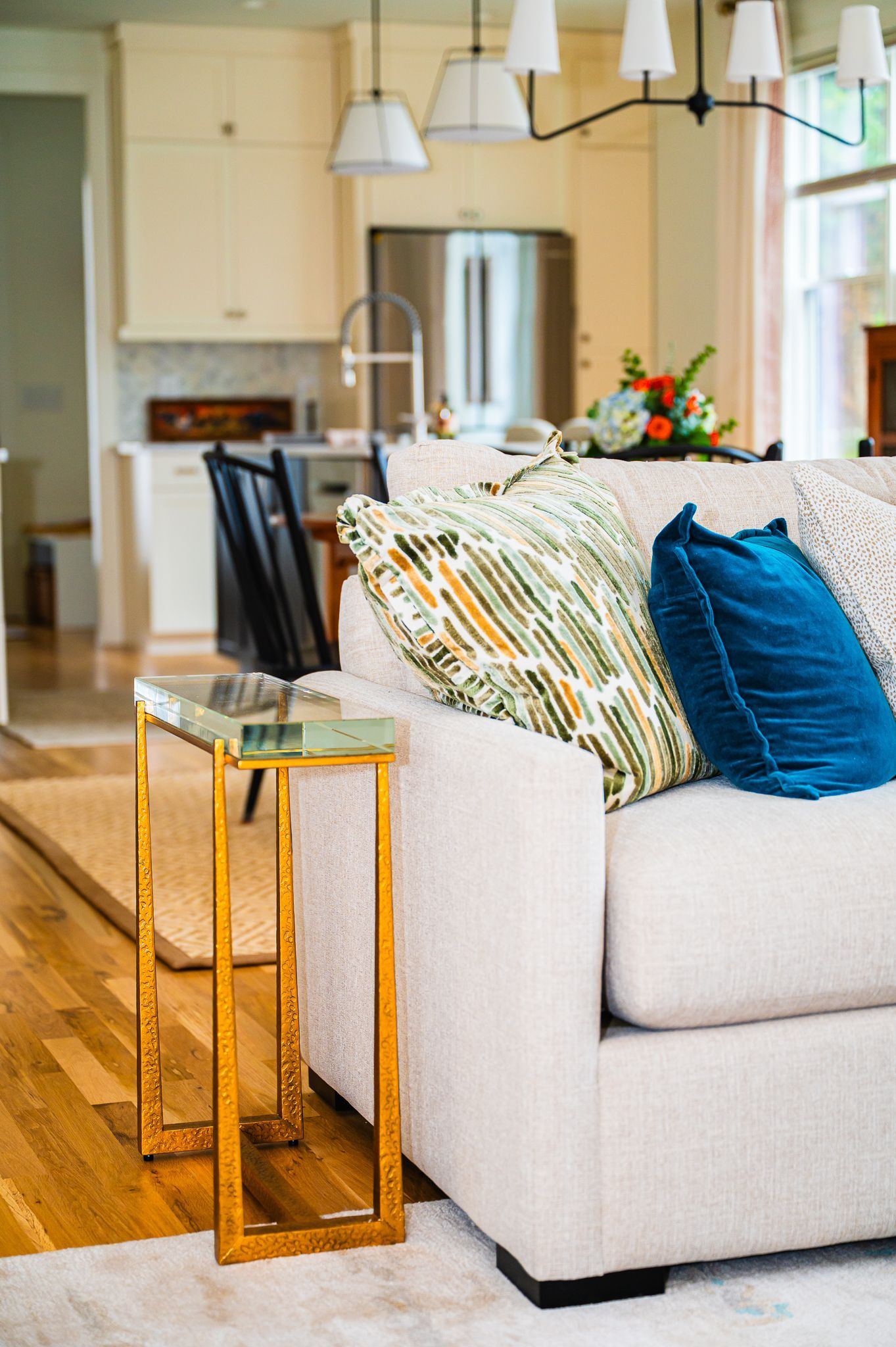 Sofa arm with green patterned and teal pillows beside two hammered brass nesting tables; kitchen pendants in background.