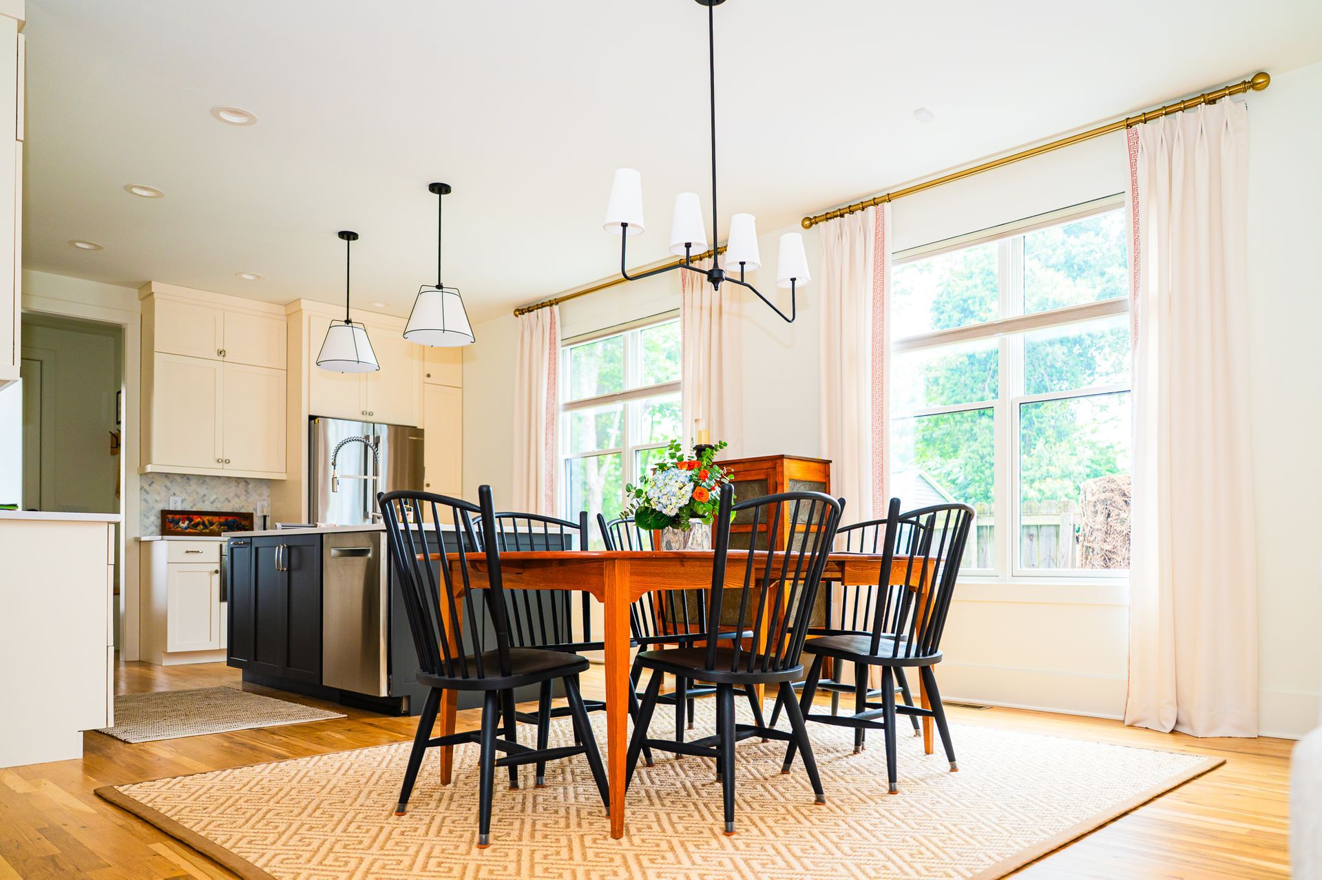 Wide view of dining table with black chairs beside a bright kitchen with cream cabinets, black island, and large windows with pale drapes.