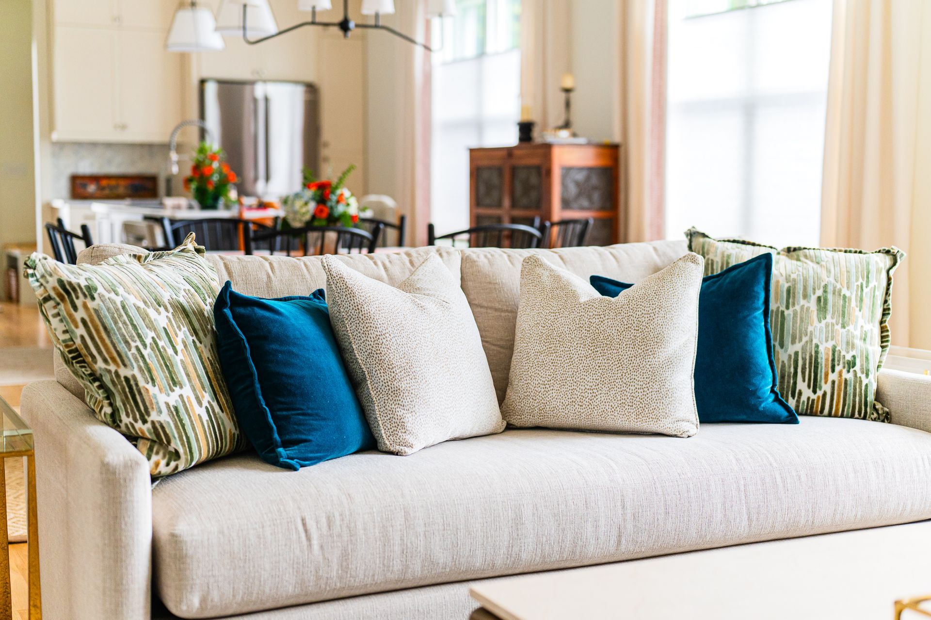 Sofa vignette with cream upholstery, teal velvet and sage patterned pillows; light, open kitchen beyond.