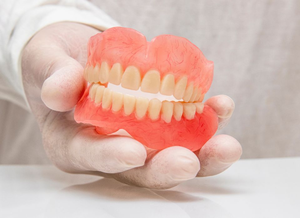 A gloved hand holding a set of upper and lower dentures, pink gums, and yellow teeth.