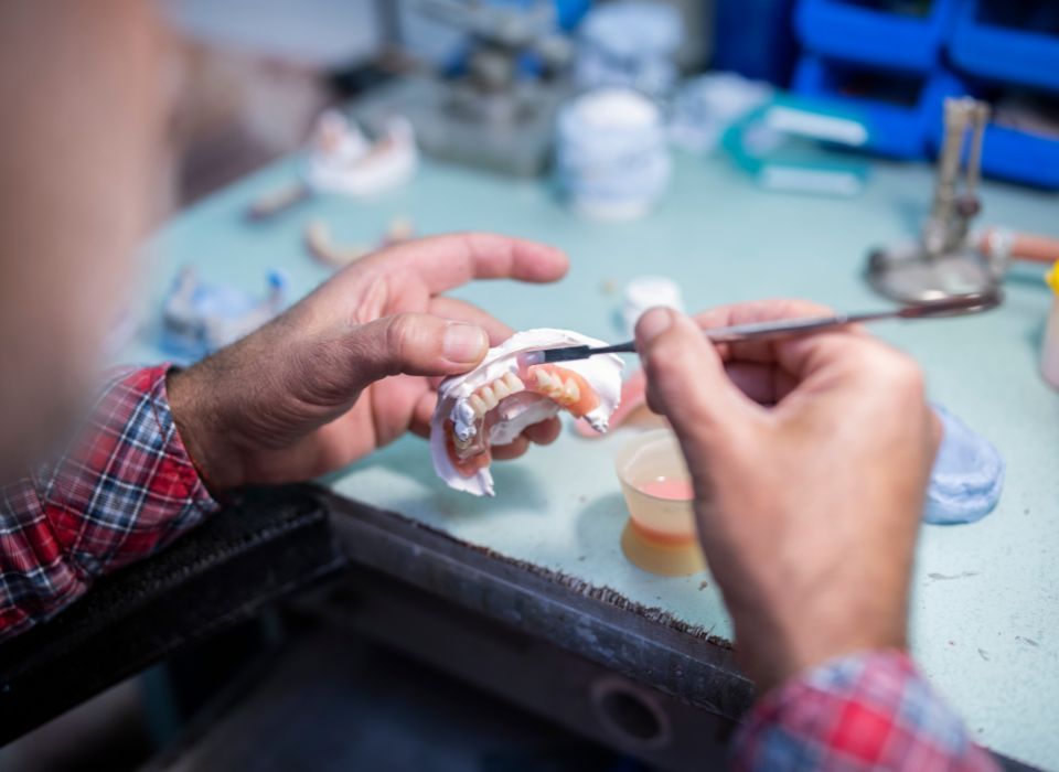 Person crafting dentures with a brush at a dental lab, wearing a plaid shirt.
