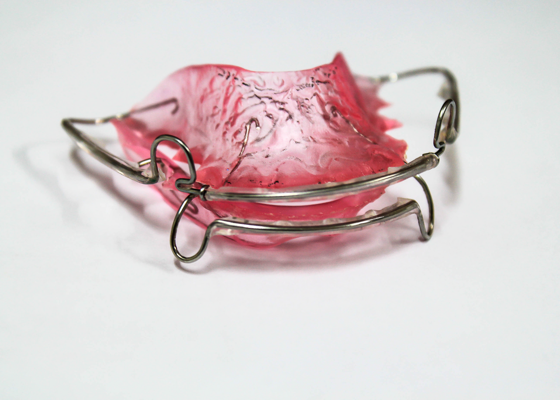 Pink and clear orthodontic retainer with metal wires, on a white surface.
