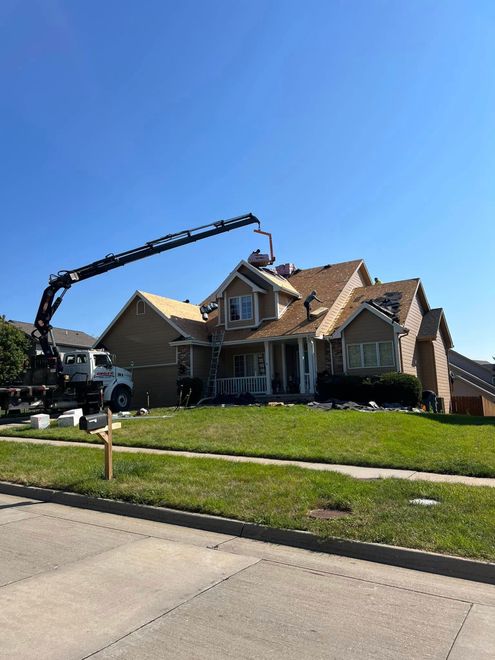 House with severe roof damage and a crane lifting debris beside it on a suburban street