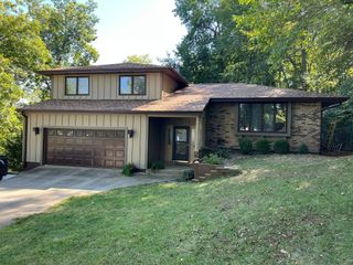 Two-story beige house with brown garage and front lawn, surrounded by trees