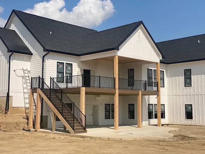 Two-story white house with black roof, front porch, staircase, and unfinished dirt yard under a blue sky