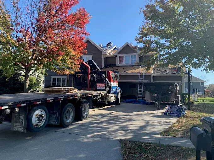 Flatbed truck unloading construction equipment in a suburban driveway beside a two-story house.