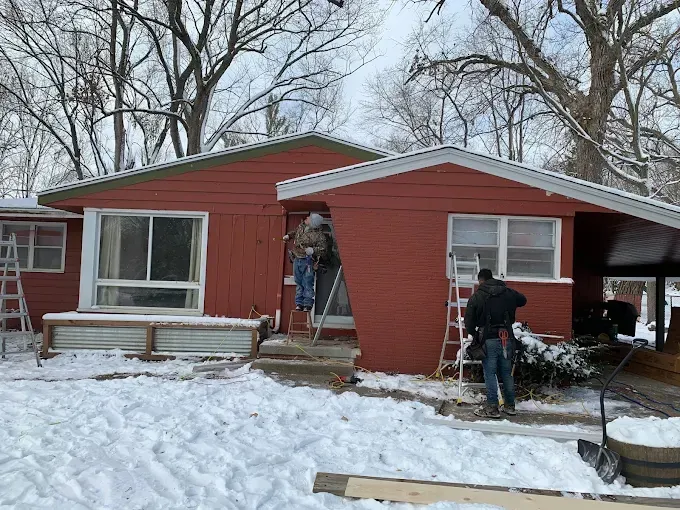 Red house with snow-covered yard; two people work at the entrance during winter.