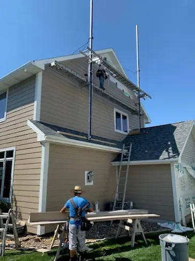 Workers repairing the roof and siding of a tan two-story house with scaffolding under a clear blue sky