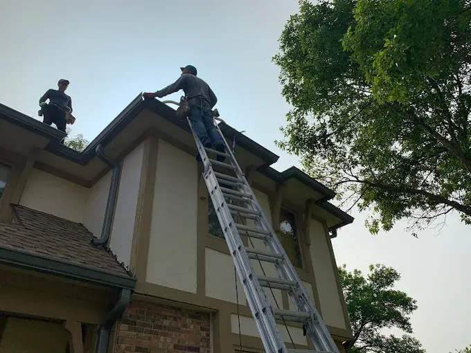 Two workers on a roof using a ladder beside a house with trees nearby