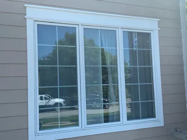 White-framed bay window on a gray house exterior with reflections of trees and cars.